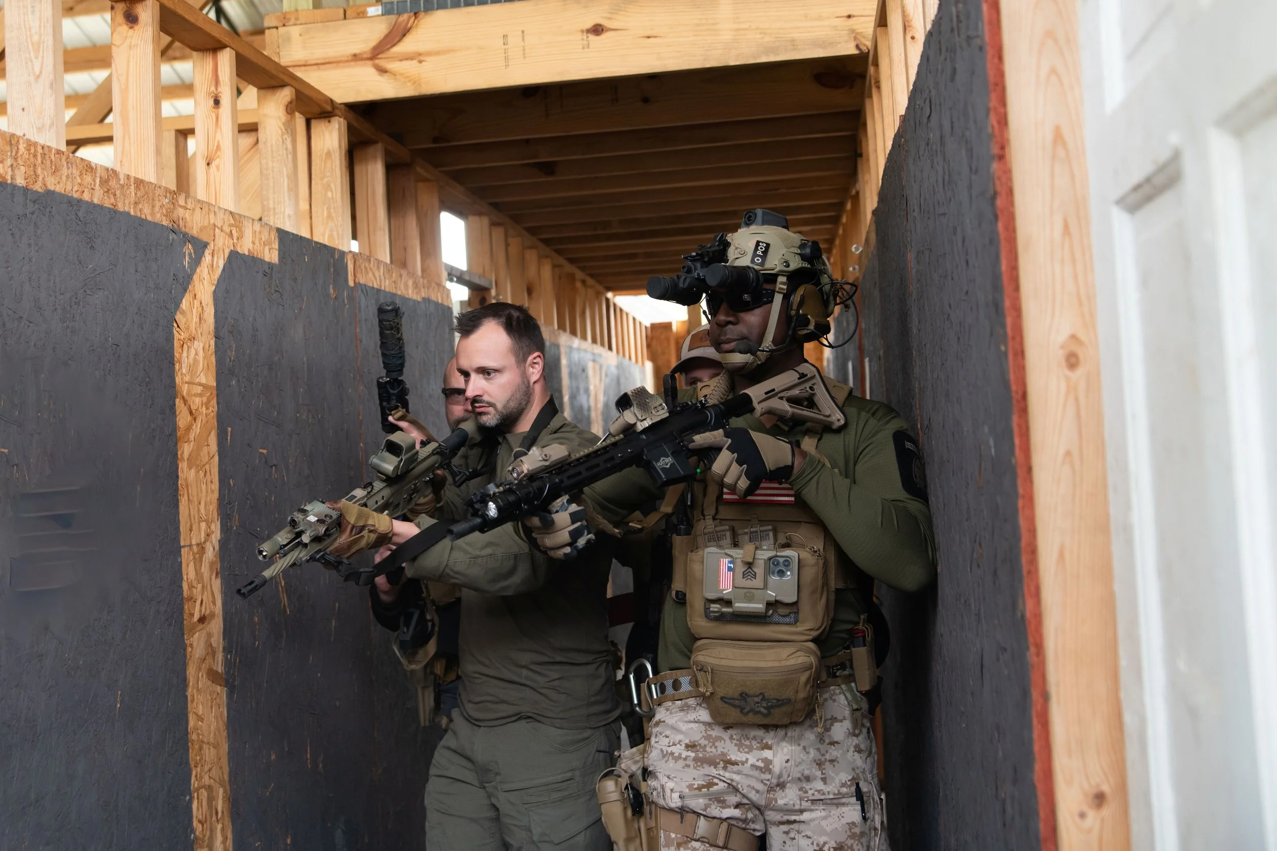 Military personnel practicing close-quarters combat in a wooden structure with black walls.
