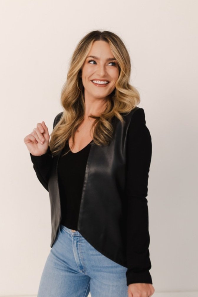 A woman with blonde wavy hair smiling while wearing a black leather vest over a black top and light blue jeans, standing against a plain white background.