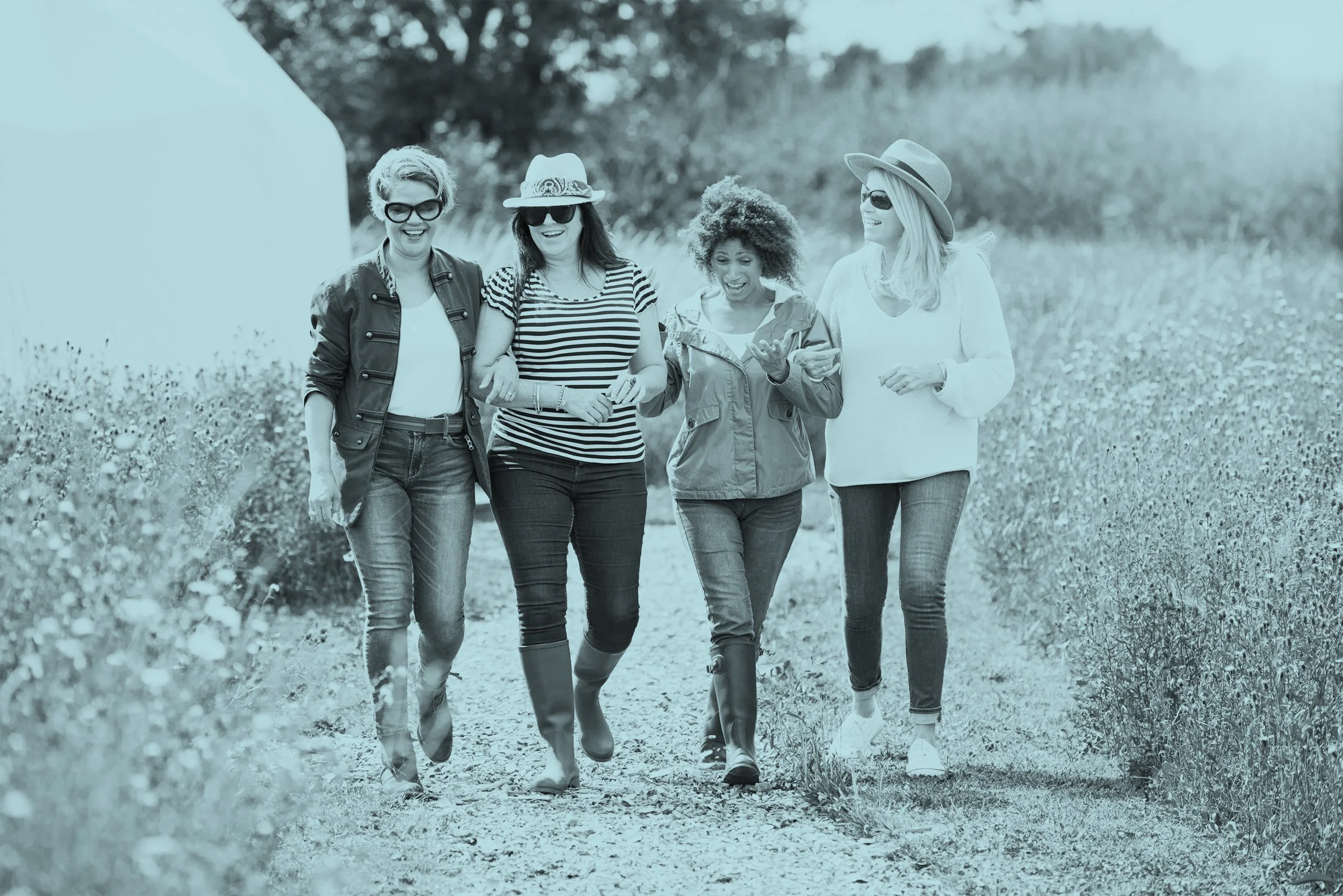 Four women walking and laughing as they stroll along a path on sunlit farmlands.