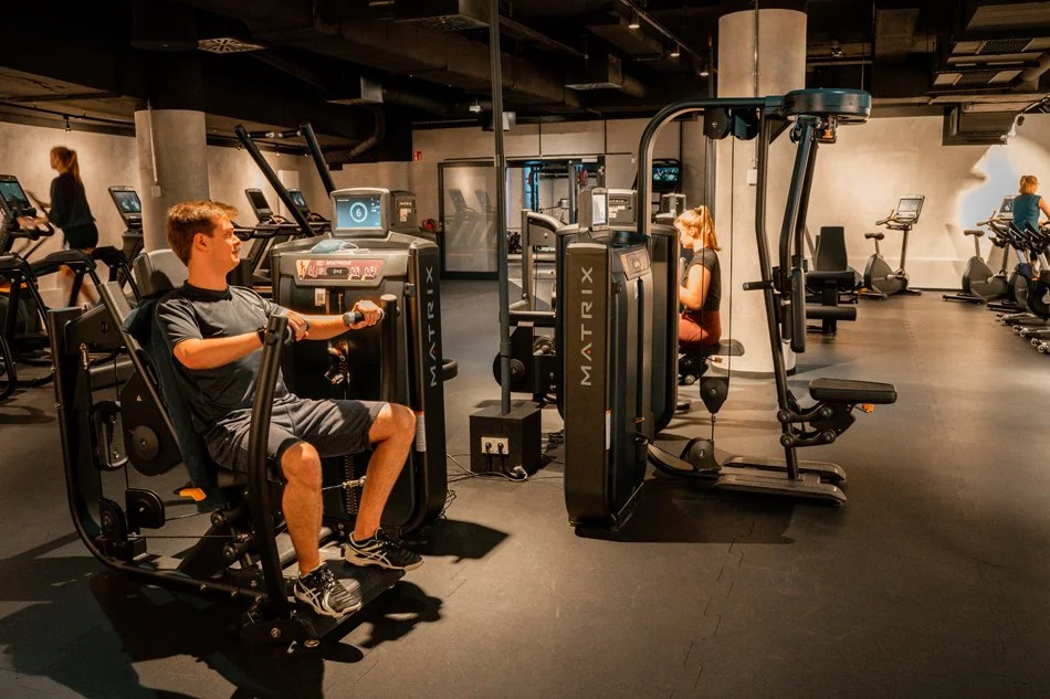 Menschen trainieren auf verschiedenen Fitnessgeräten in einem modernen Fitnessstudio, darunter ein Mann auf einem Sitzergometer und eine Frau auf einem Rudergerät.