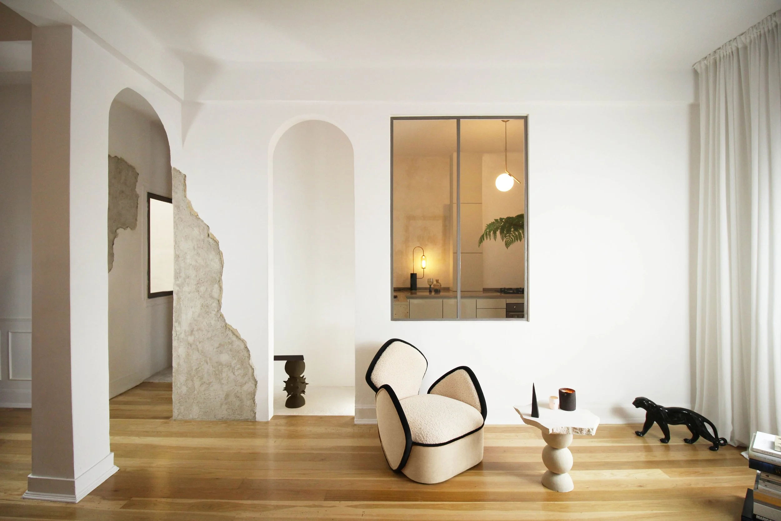 Modern living room with a white armchair, small side table, black panther statue, and decorative objects on the floor, with a large window covered by white curtains, and a kitchen visible through a window in the wall.