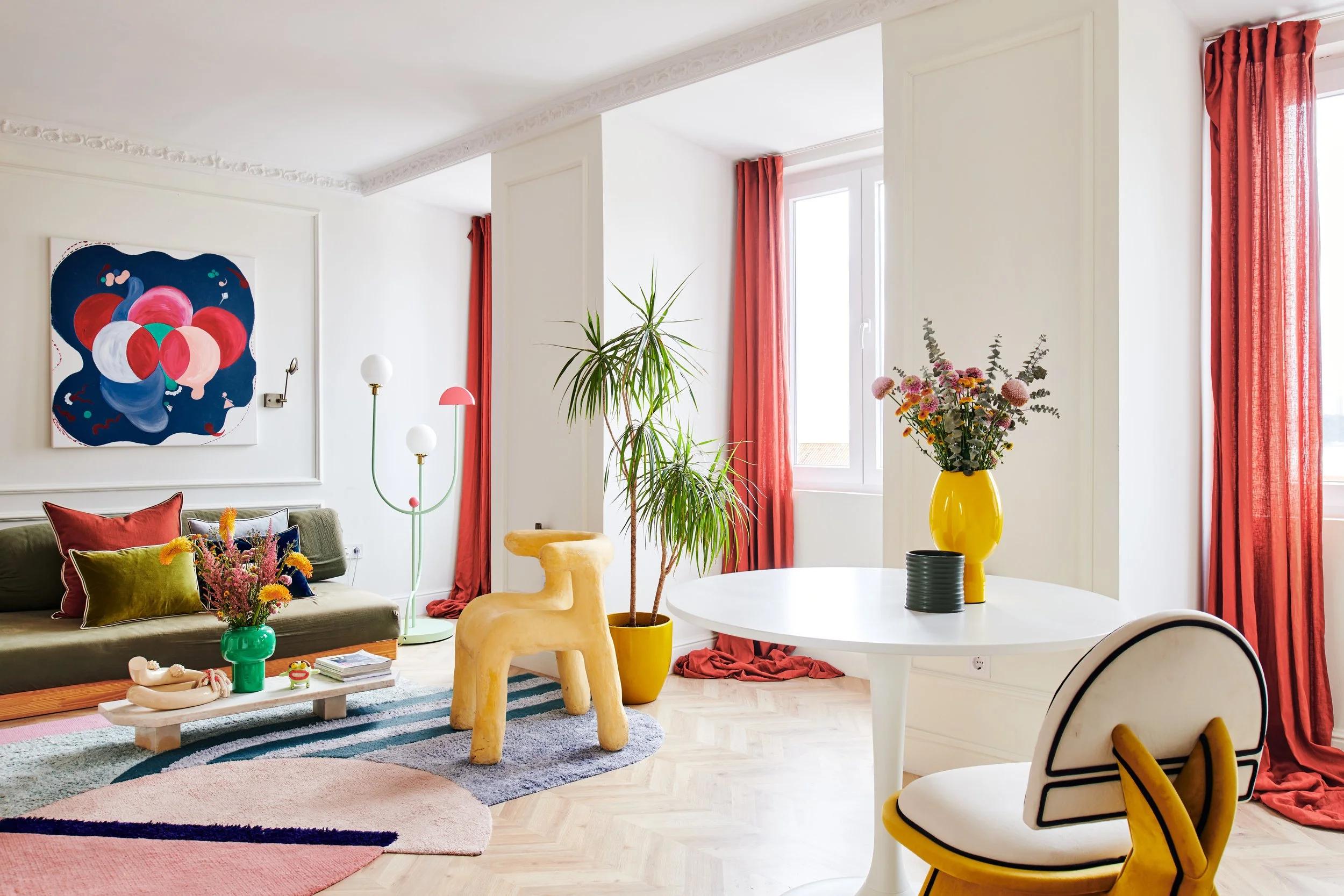 Living room with white walls, red curtains, a green sofa with colorful pillows, a white round table with a yellow vase of pink and purple flowers, a cream-colored sculptural chair, potted plants, and artwork on the wall.