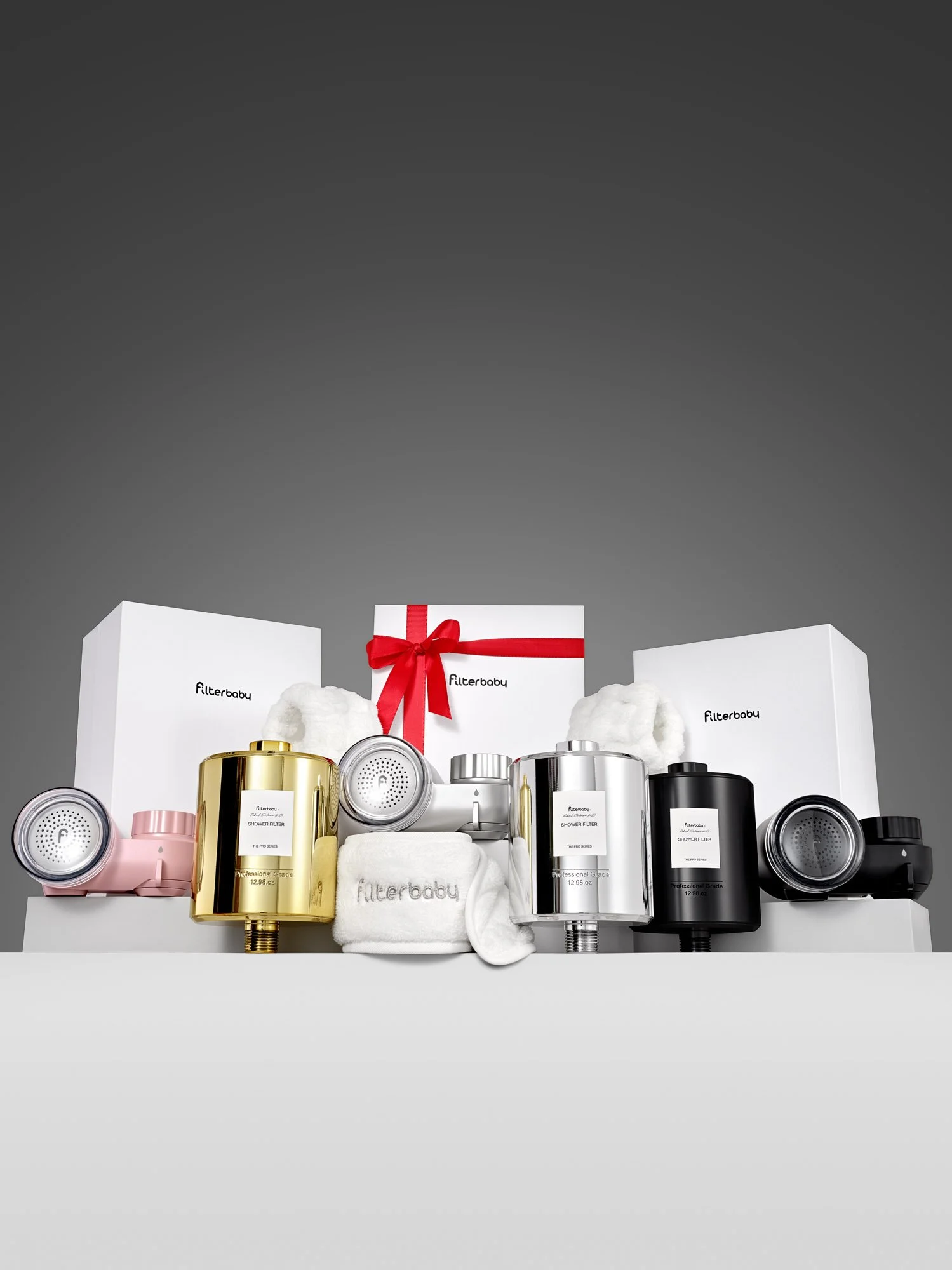 Large family bundle image showing 3 Filterbaby Pro Series Shower Filters in gold, silver, and black, paired with 3 Filterbaby Skincare Faucet Water Filters in pink, silver, and black, a branded headband and cuffs, and 3 branded boxes. The middle box 