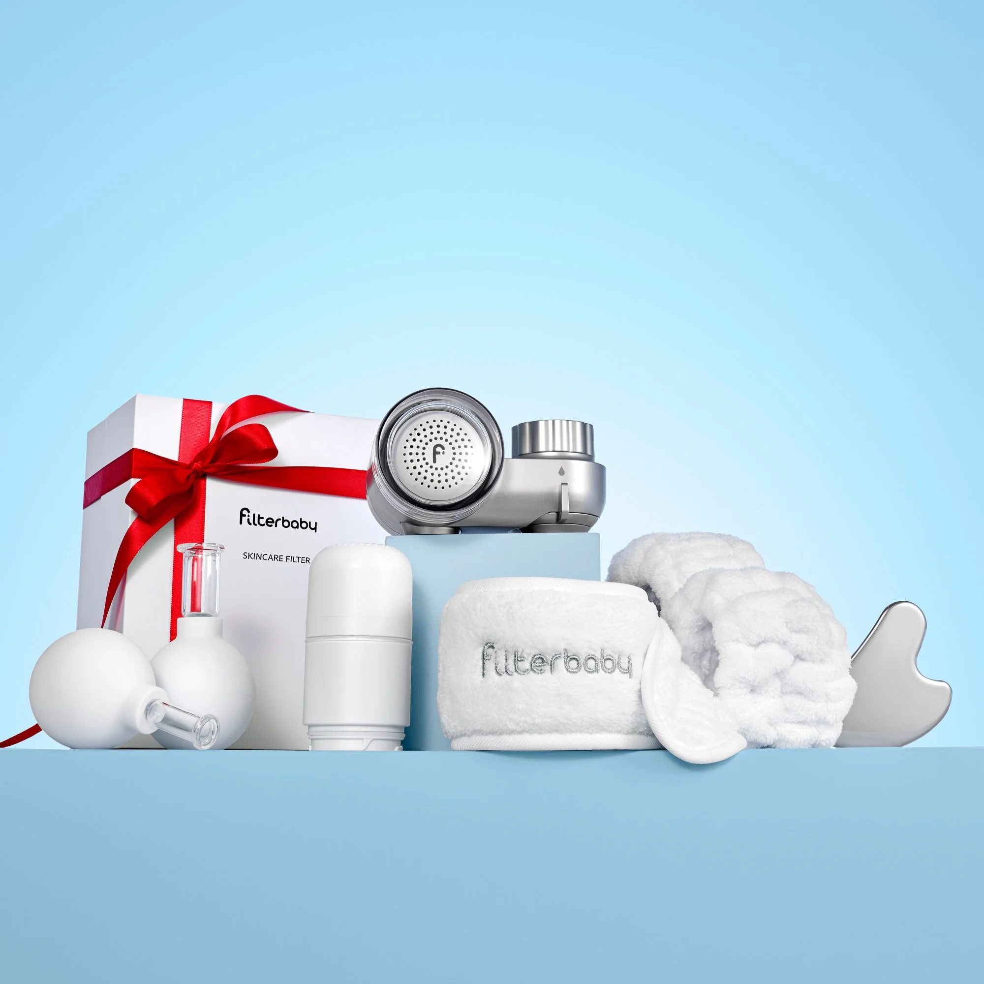 Bundle image of the Filterbaby Skincare Faucet Water Filter in silver along with several gifts with purchase, including a branded headband and cuffs, metal silver gua sha, replacement filter, and facial cupping tools. The filterbaby is on a riser box
