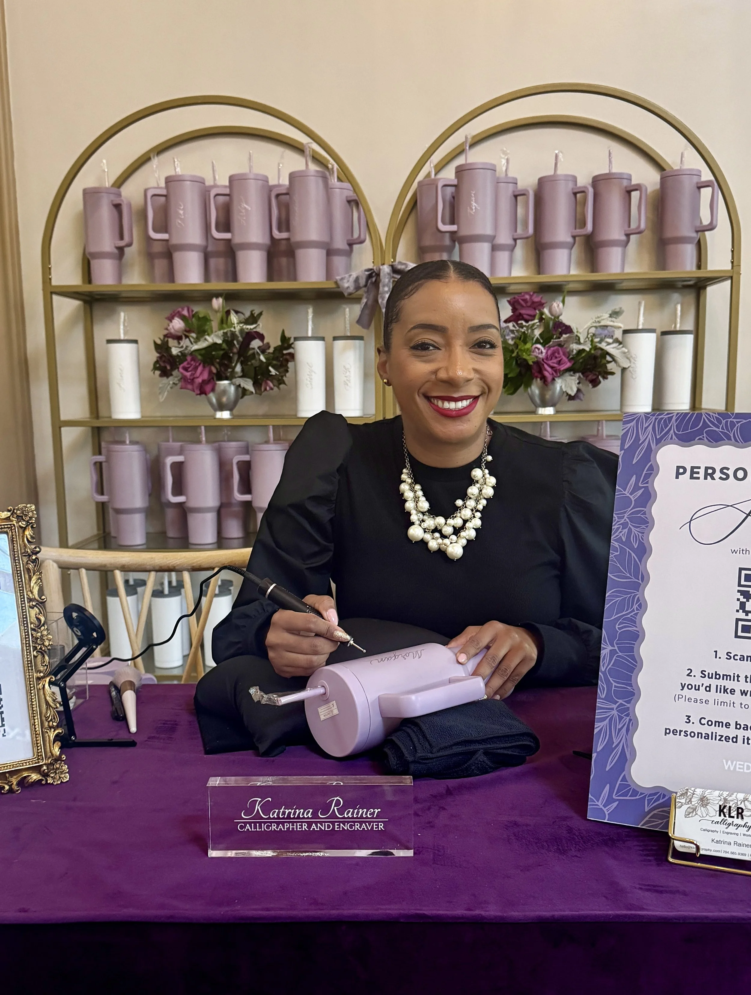 Woman smiling at a desk with champagne, high heel-shaped bottle, and writing on perfume bottle.