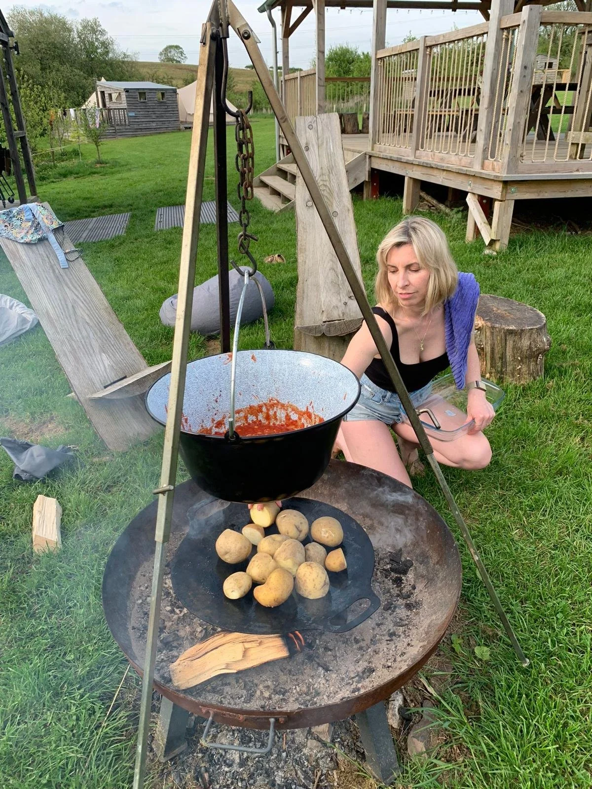 A woman cooking potatoes over an open fire pit outdoors, with a large cooking pot hanging over the fire and potatoes on a flat stone in the fire.