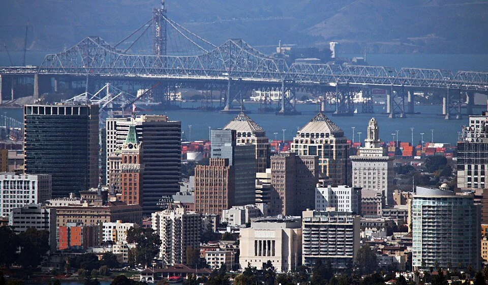 City skyline of Oakland, CA with tall buildings and the Bay Bridge in the background.