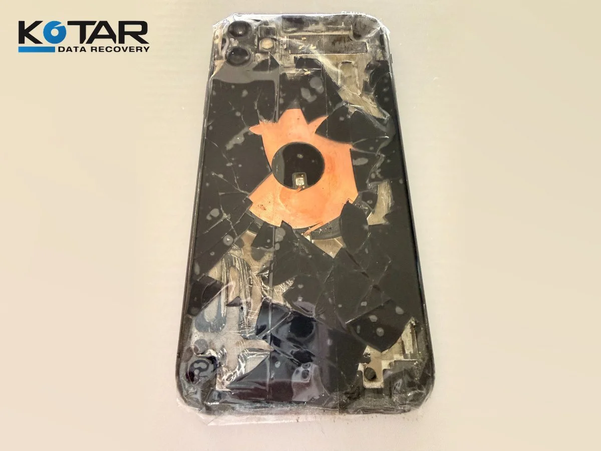 Severely damaged smartphone with cracked black rear glass, exposed copper-colored circular component, and Kotar Data Recovery logo in the upper-left corner.