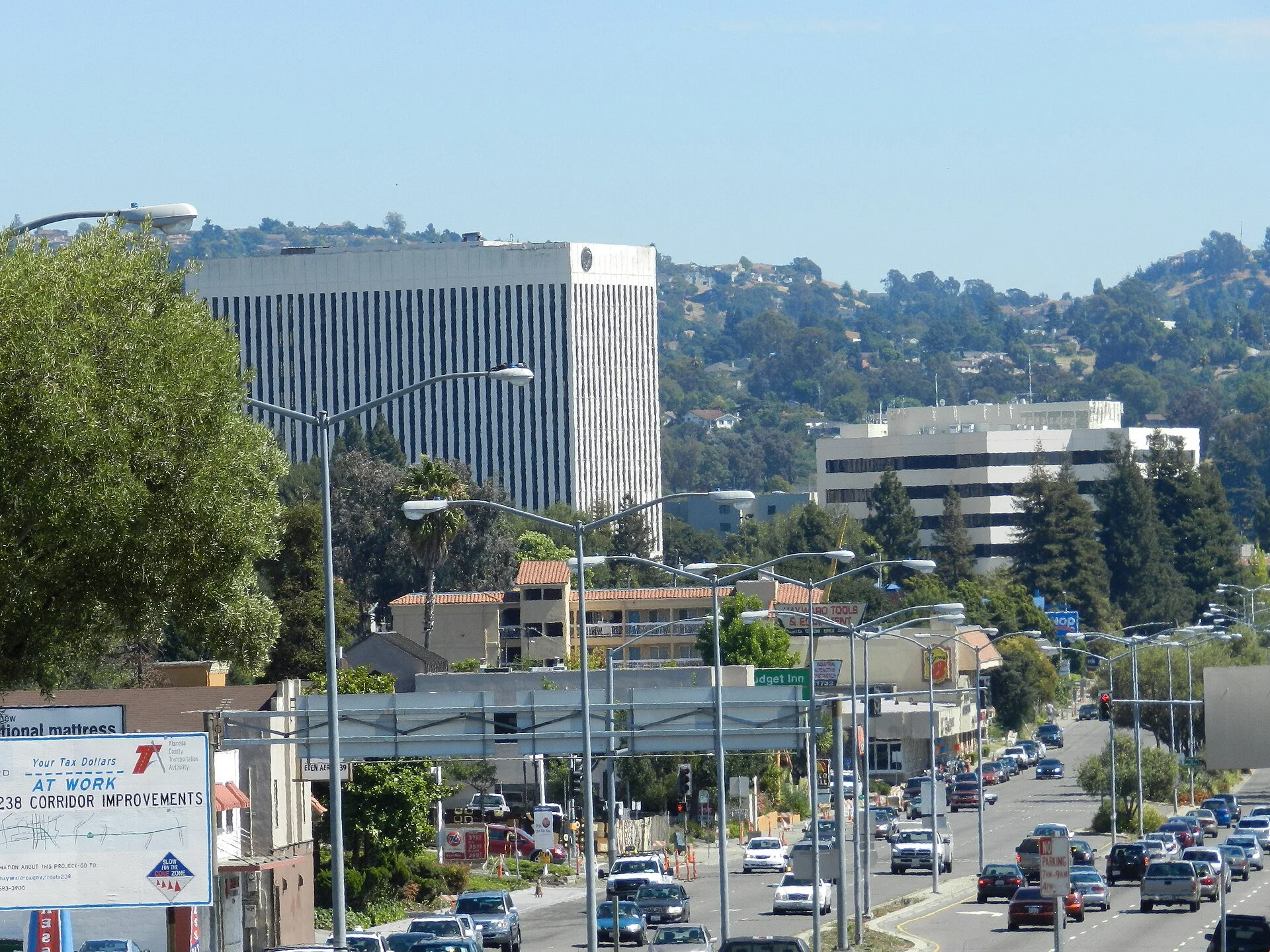 City street in Hayward, CA with cars, streetlights, commercial buildings, and taller office buildings in the background on a sunny day.