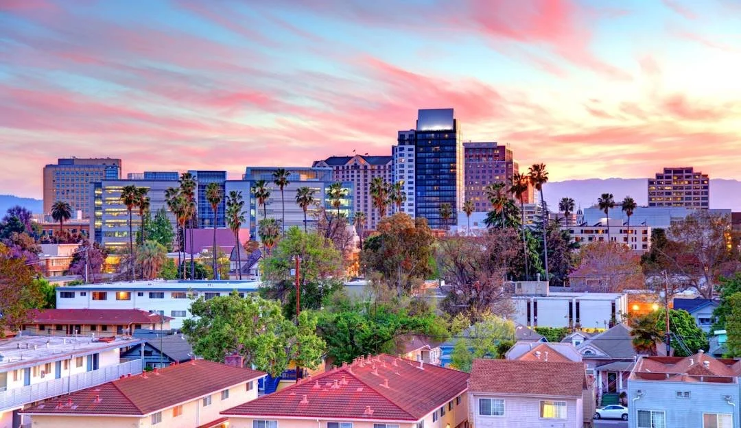 City skyline of San Jose, CA at sunset with colorful sky, tall buildings, palm trees, and residential houses in the foreground.