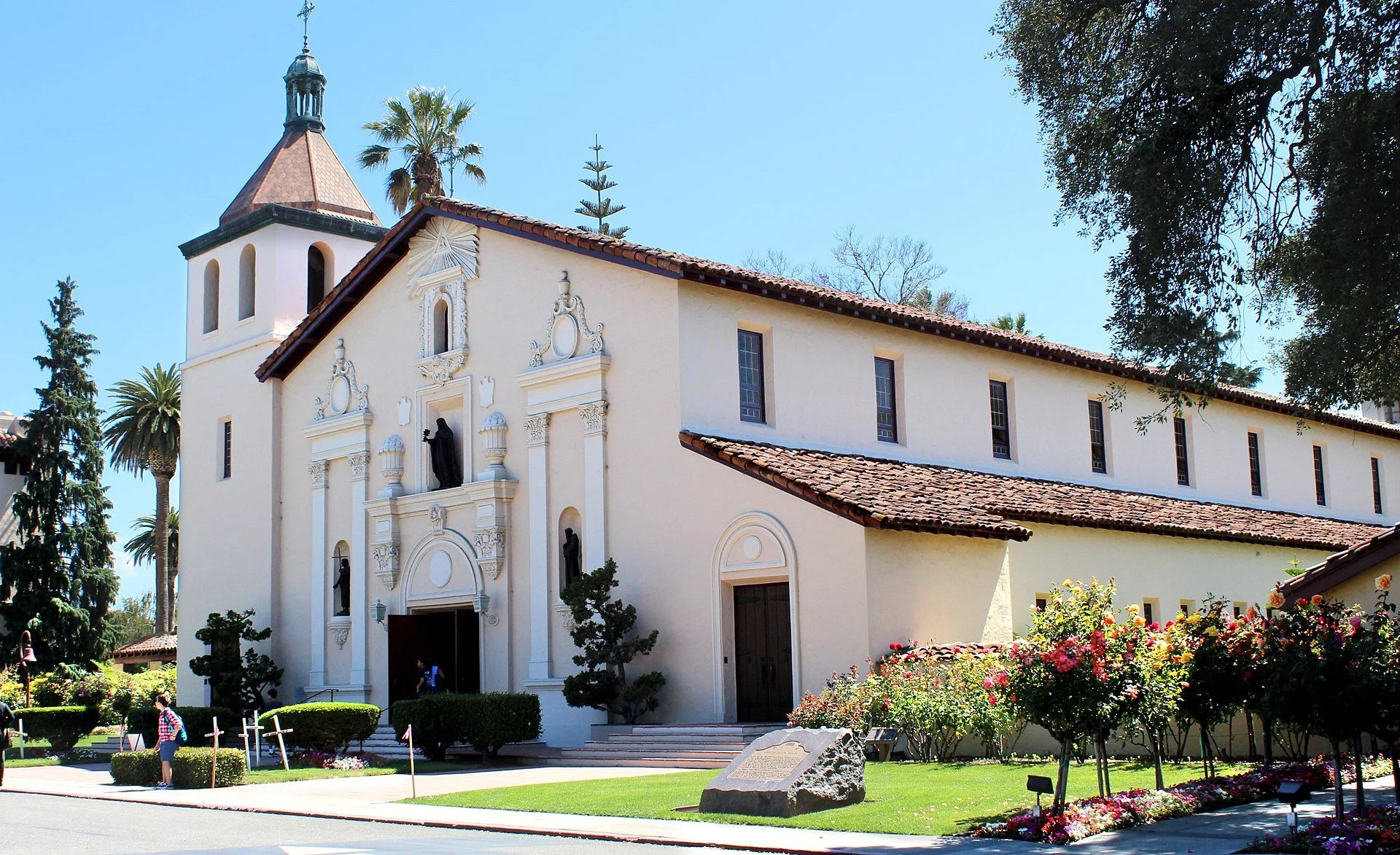 White church in Santa Clara, CA with bronze tiled roof, decorative sculptures, and a small bell tower, surrounded by palm trees and flowering shrubs.