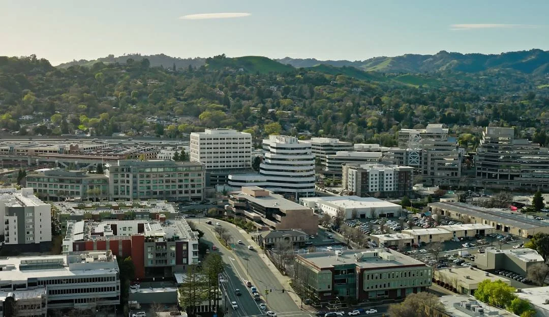 A cityscape of Walnut Creek, CA with modern buildings, parking lots, and nearby green hills.