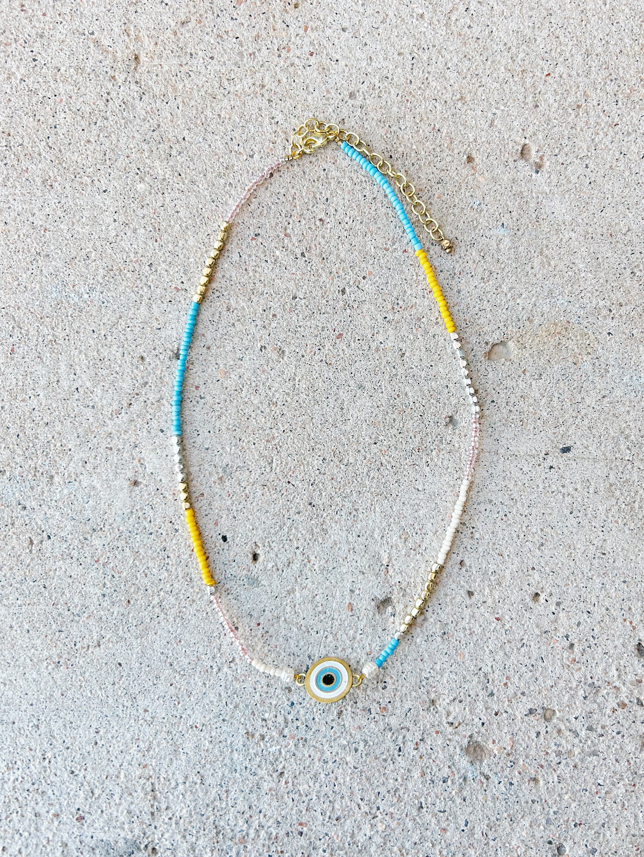 The Double Vision Necklace features seed beads in sky blue, pink, mustard yellow, white, and gold. There is a small eyeball charm in the center of the necklace.