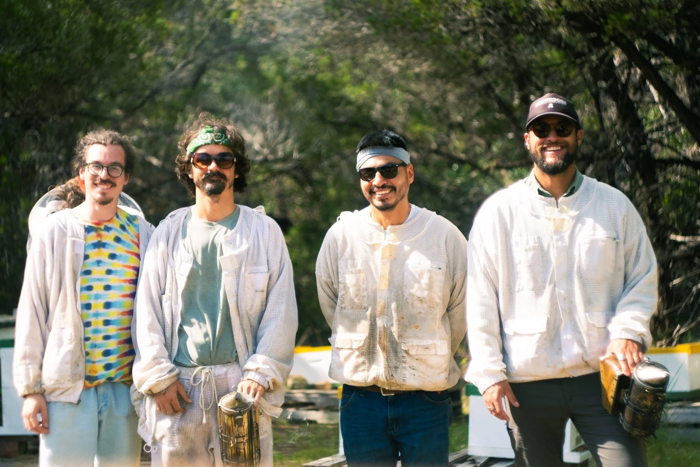 Four beekeepers smiling at the camera, with some holding smokers, dressed in beekeeping protective gear.