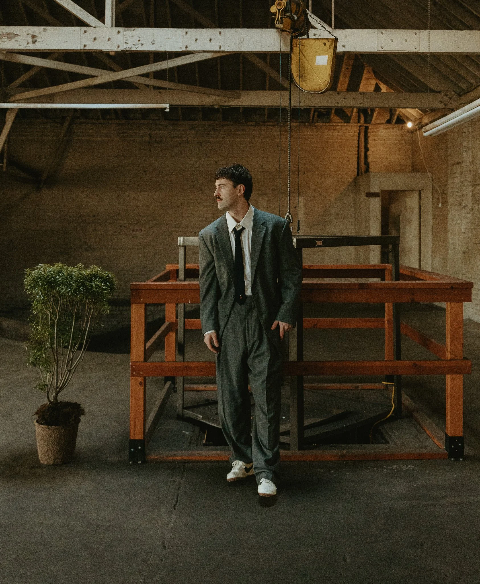 A man in a gray suit with a tie stands in an industrial-style room with exposed brick walls and wooden structural beams. He is looking to his left, with his right hand in his pocket, while a potted plant is on his left on the floor. Behind him is a large wooden platform with a black metal frame and equipment hanging from the ceiling.
