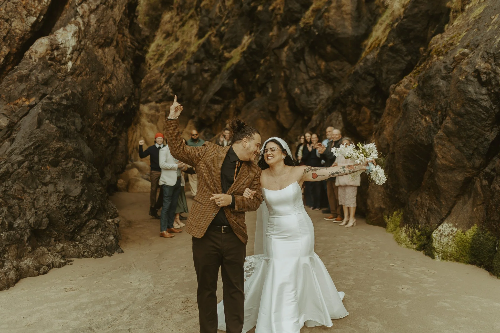 A bride and groom at their wedding in a rocky outdoor setting, with guests clapping and celebrating in the background.