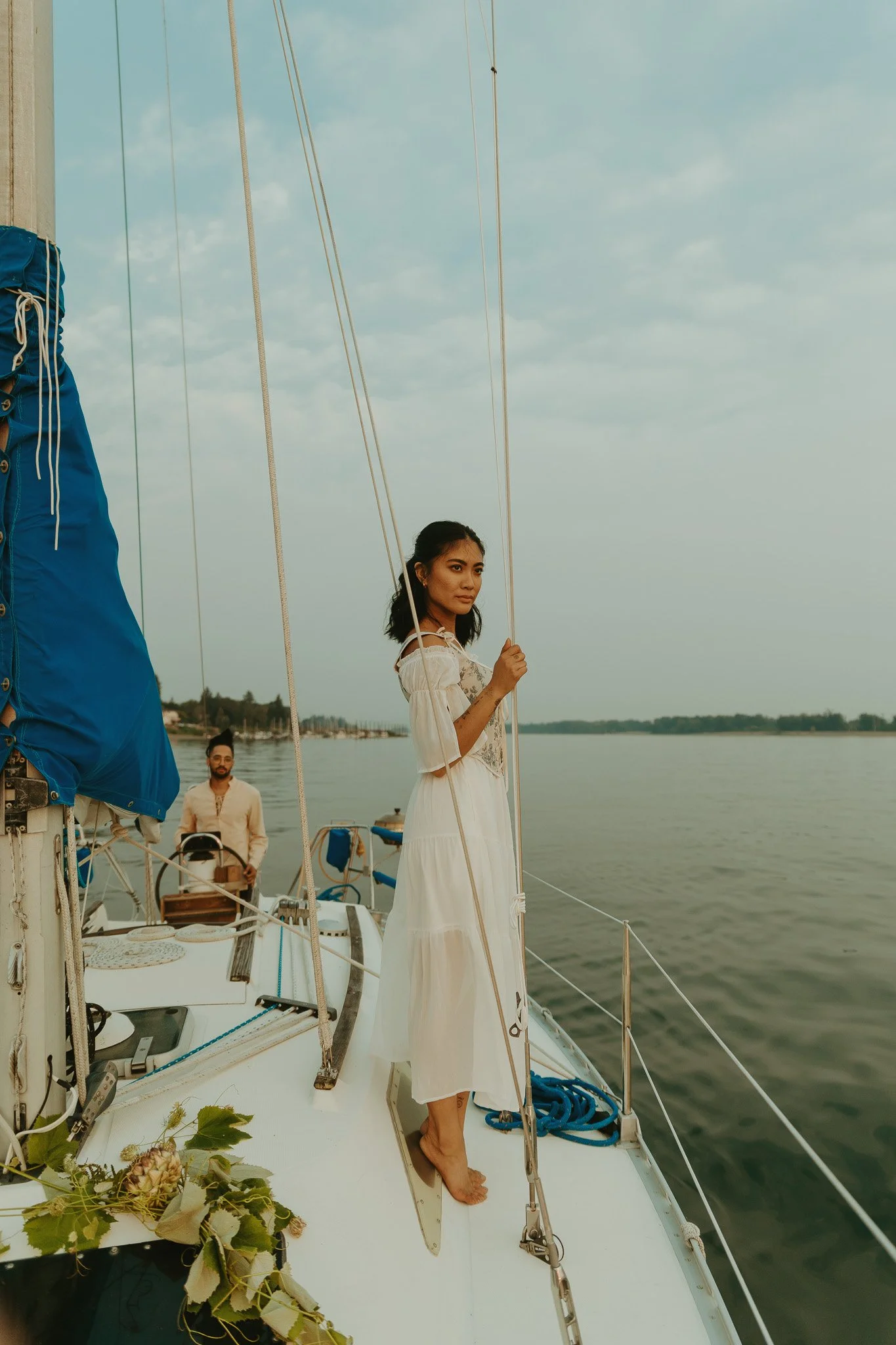 A woman in a white dress standing barefoot on a sailboat, holding a rope, with a man in the background steering the boat on a calm body of water.