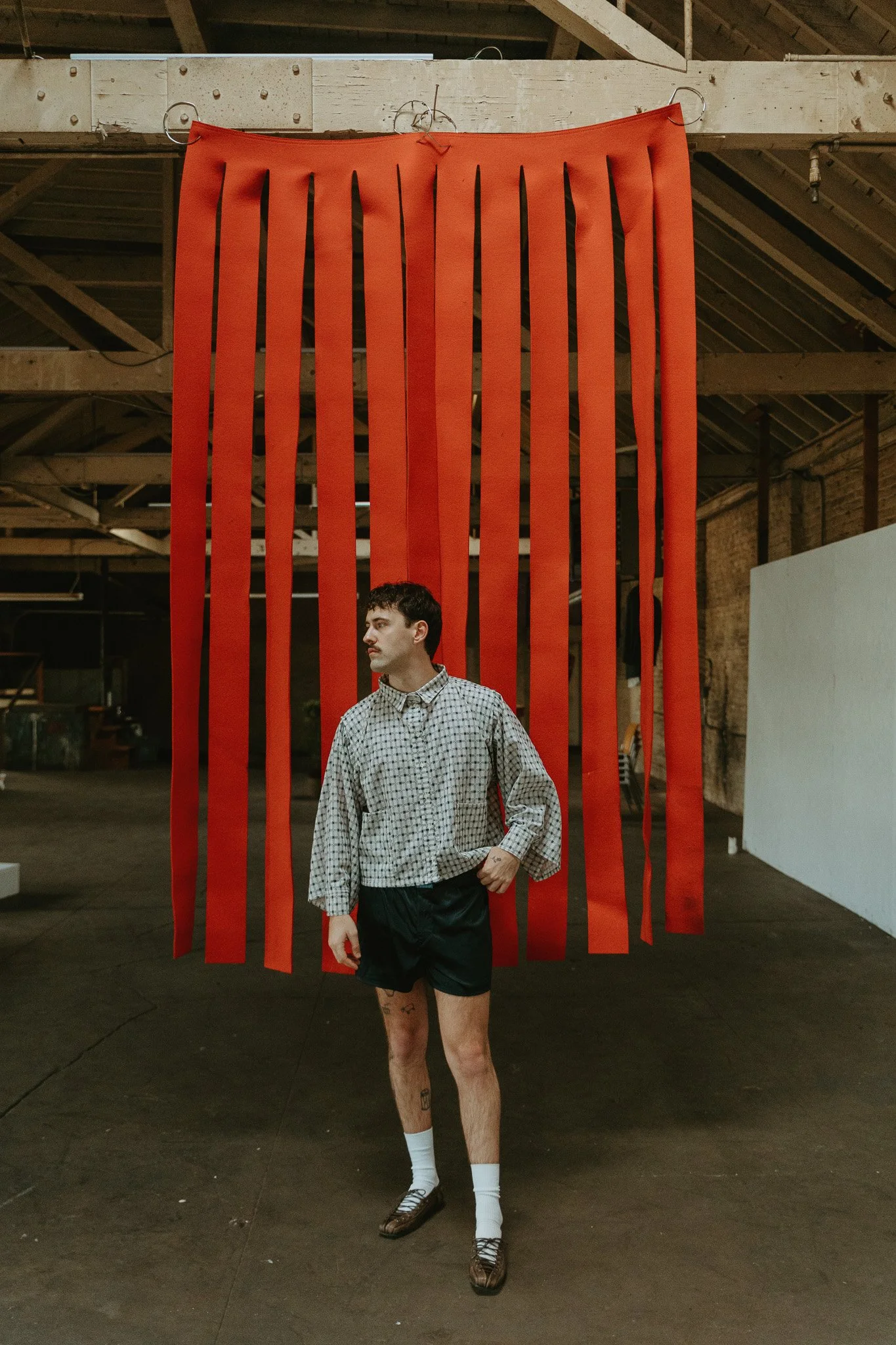 A man standing inside a spacious industrial-style room with exposed brick and wooden beams, in front of a large red hanging installation made of vertical red strips suspended from the ceiling.