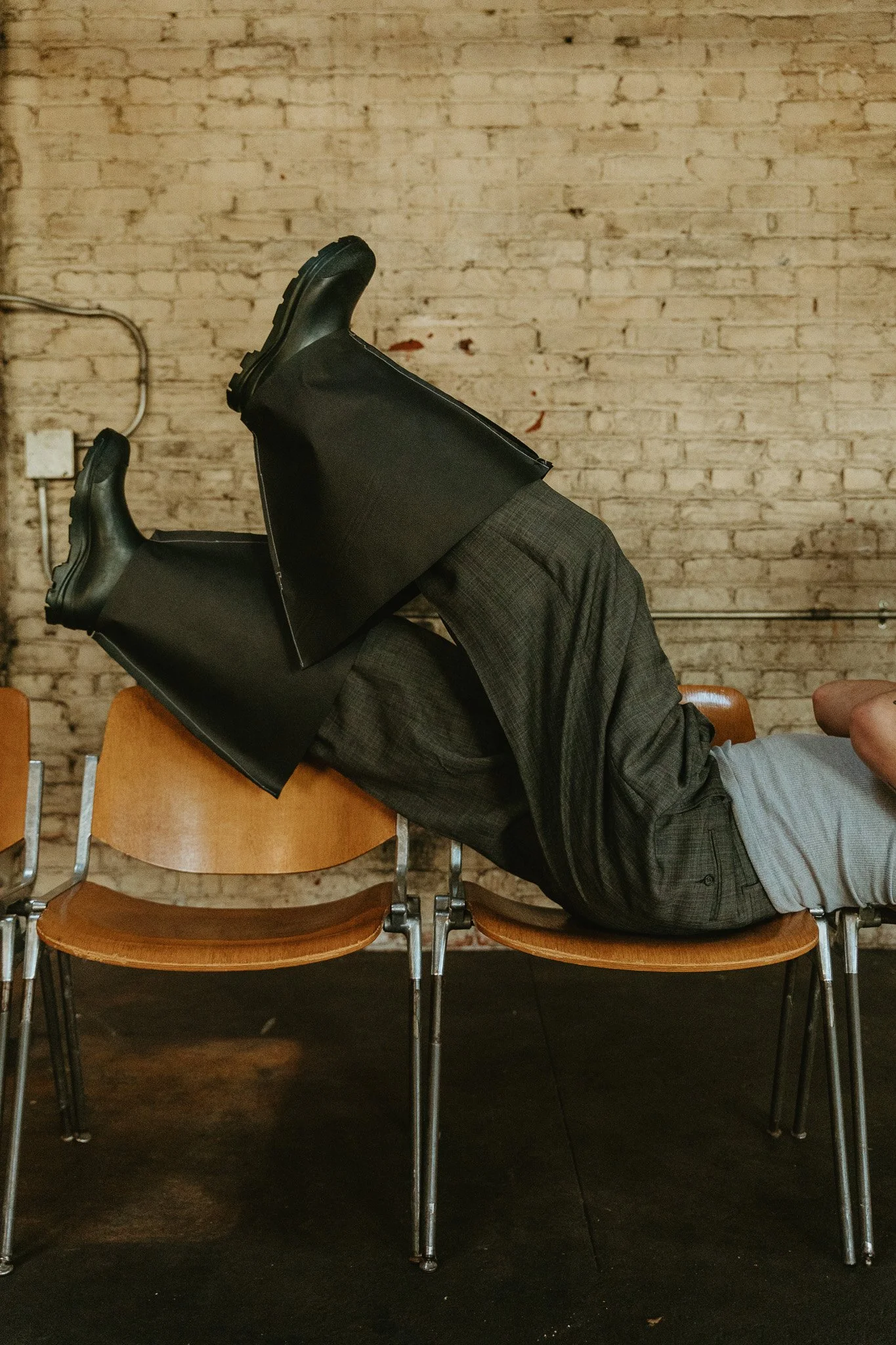 Person lying on a row of chairs with legs and feet raised above their face, wearing black shoes, grey dress pants, a grey shirt, and with their face obscured.