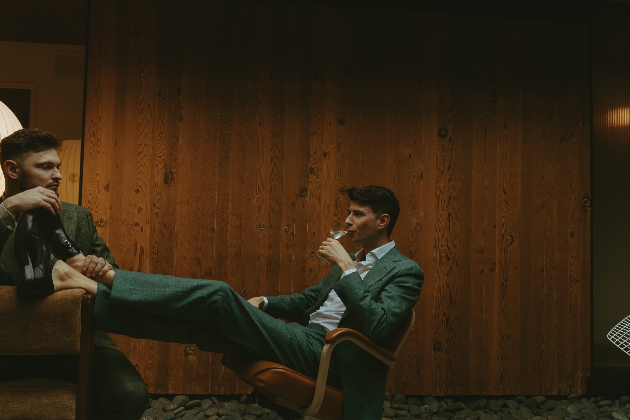 Two men in suits relaxing indoors; one man is sitting on a chair drinking a cocktail while the other man is sitting on a sofa with his foot resting on the other's lap.