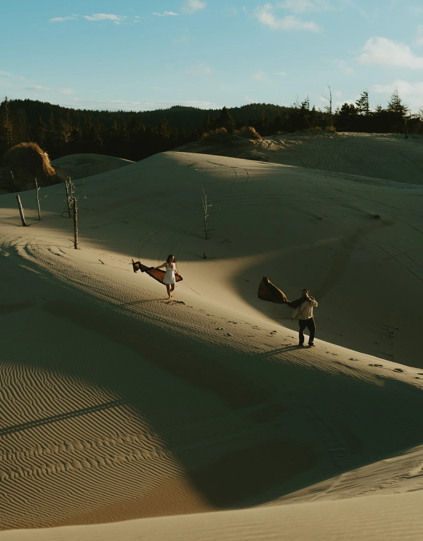 Two people walking on sand dunes, one holding a blanket, with dry trees and a forest in the background under a partly cloudy sky.