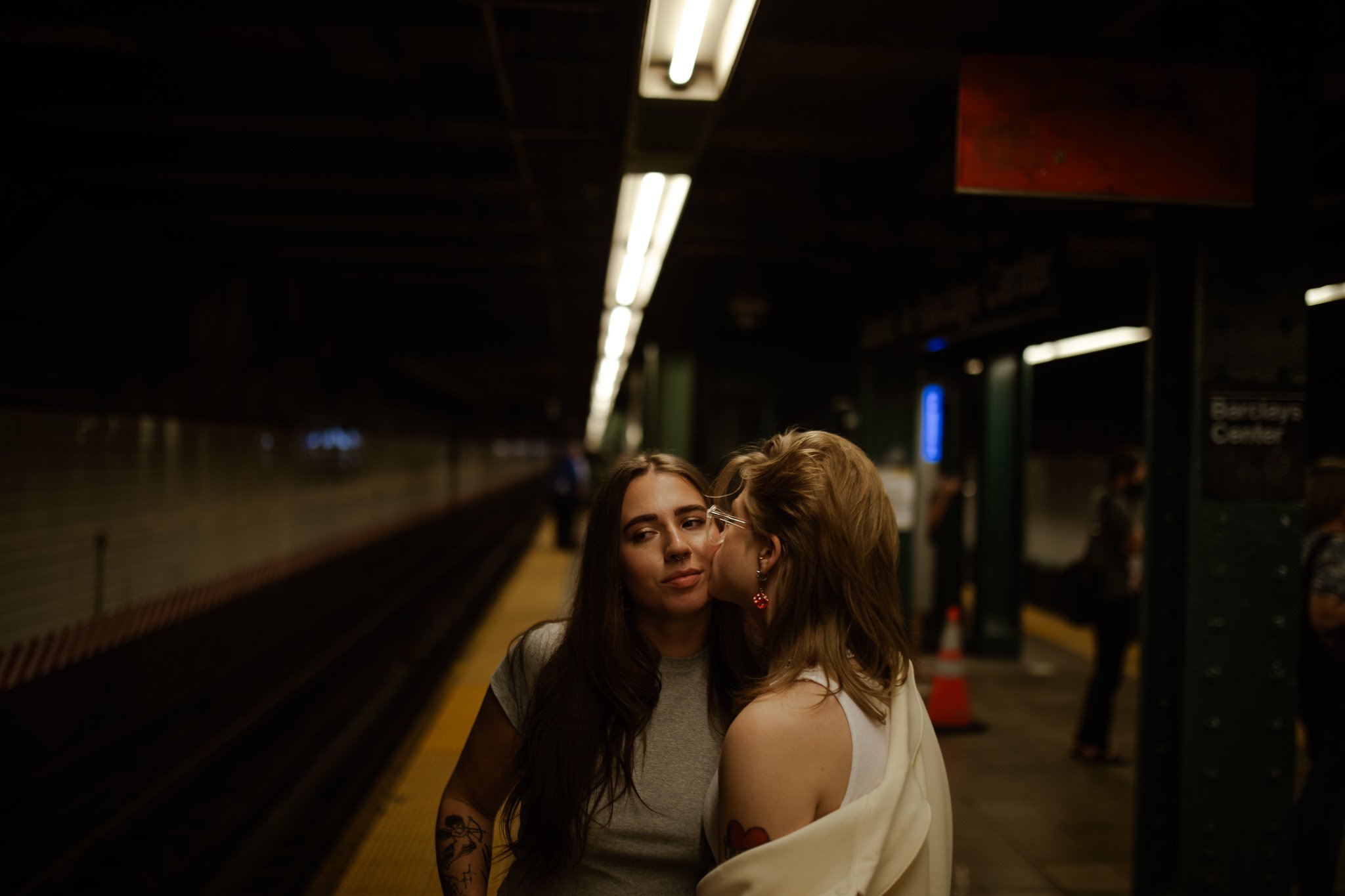 Two women standing on a subway platform, one is kissing the other on the cheek. The platform is dimly lit with a few other people in the background.