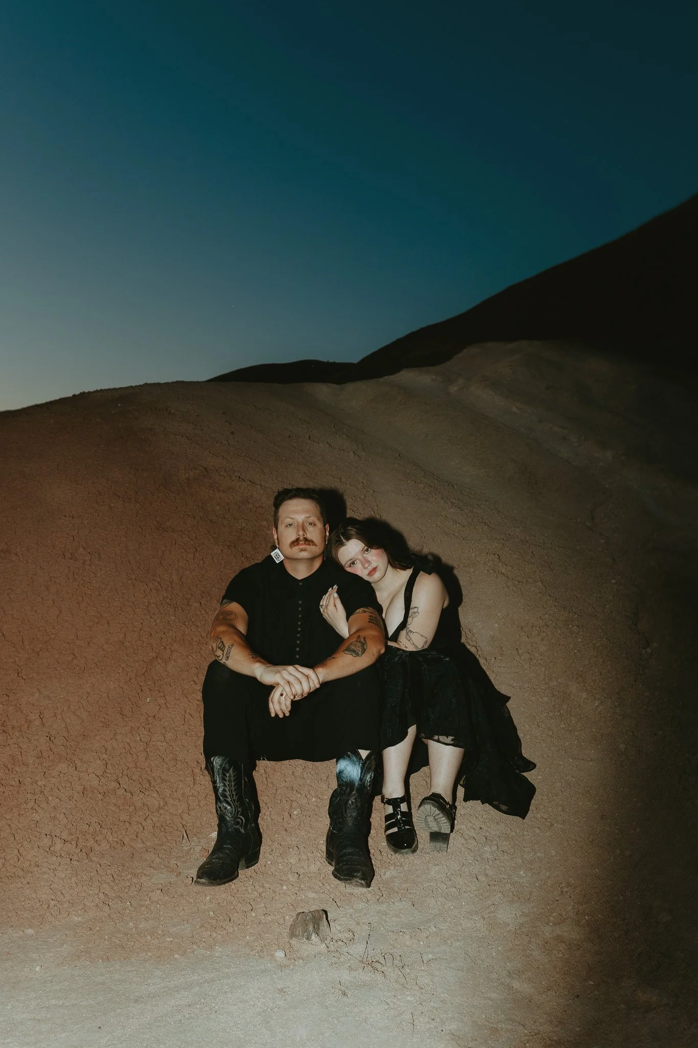 Two people sitting in a desert landscape during dusk, with hill terrain and a gradient sky in the background.