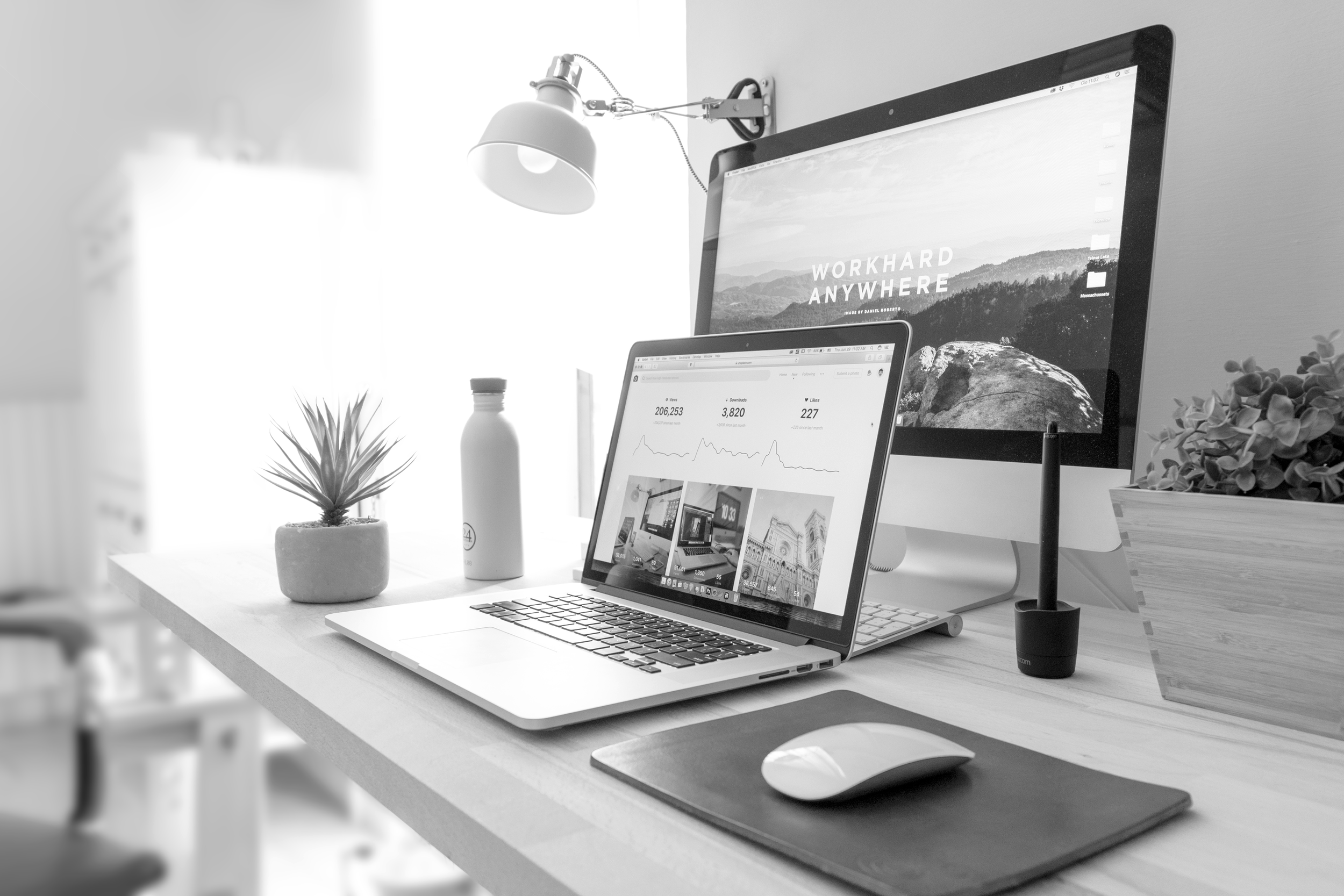 A modern workspace with a laptop, desktop monitor, mouse, desk lamp, potted plant, water bottle, and wooden box on a wooden desk.