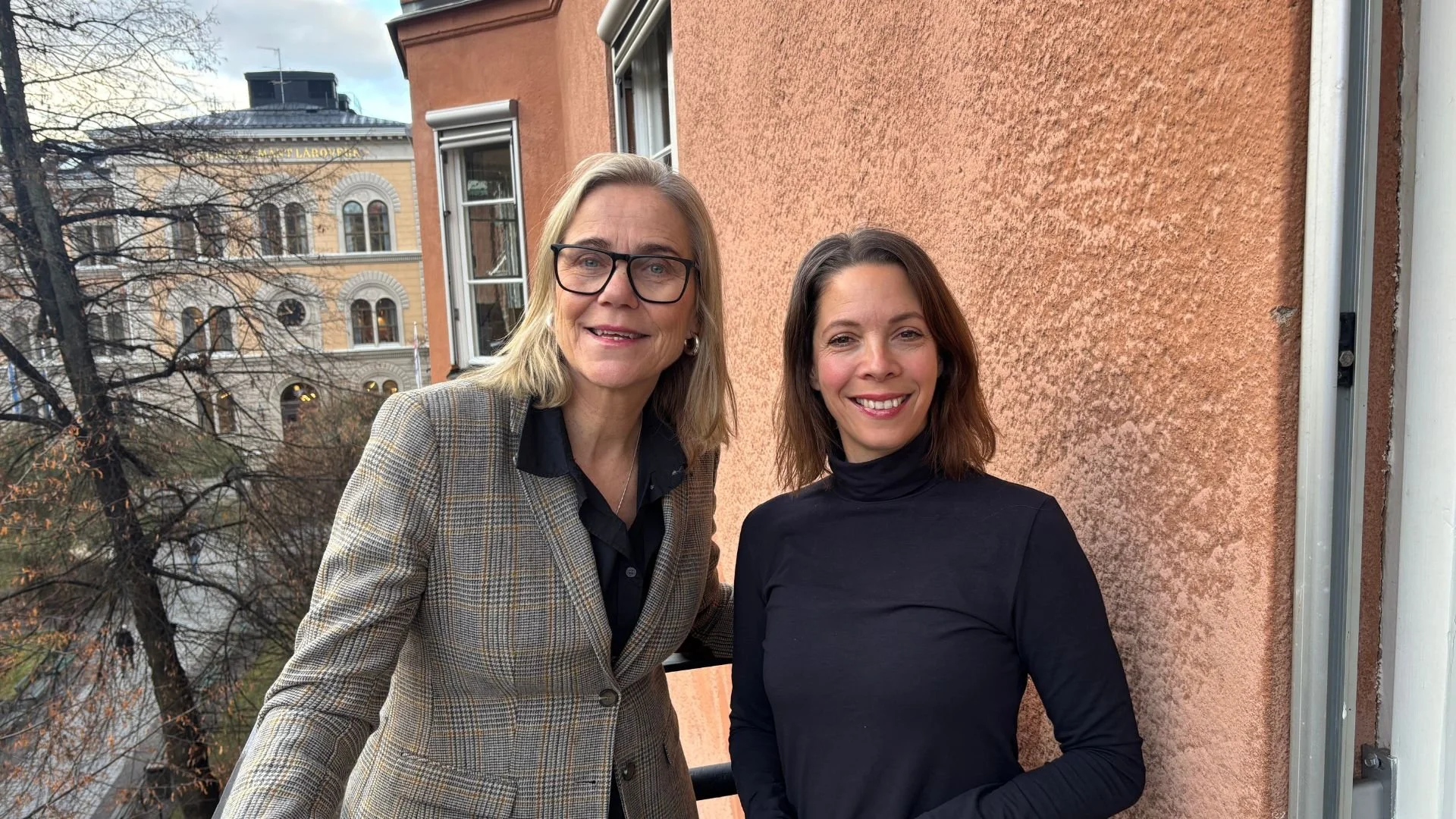 Anne-Lee och Tamara är specialister på konflikthantering i arbetslivet. Här står de bredvid varandra med en husfasad i bakgrunden och kollar leende in i kameran.