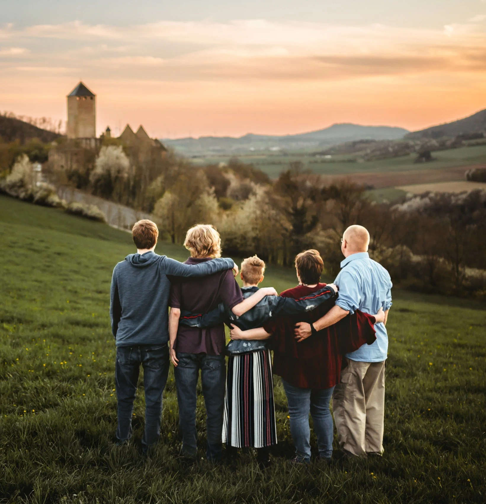 family with older kids embracing facing away from camera in beautiful field at sunset with castle in background
