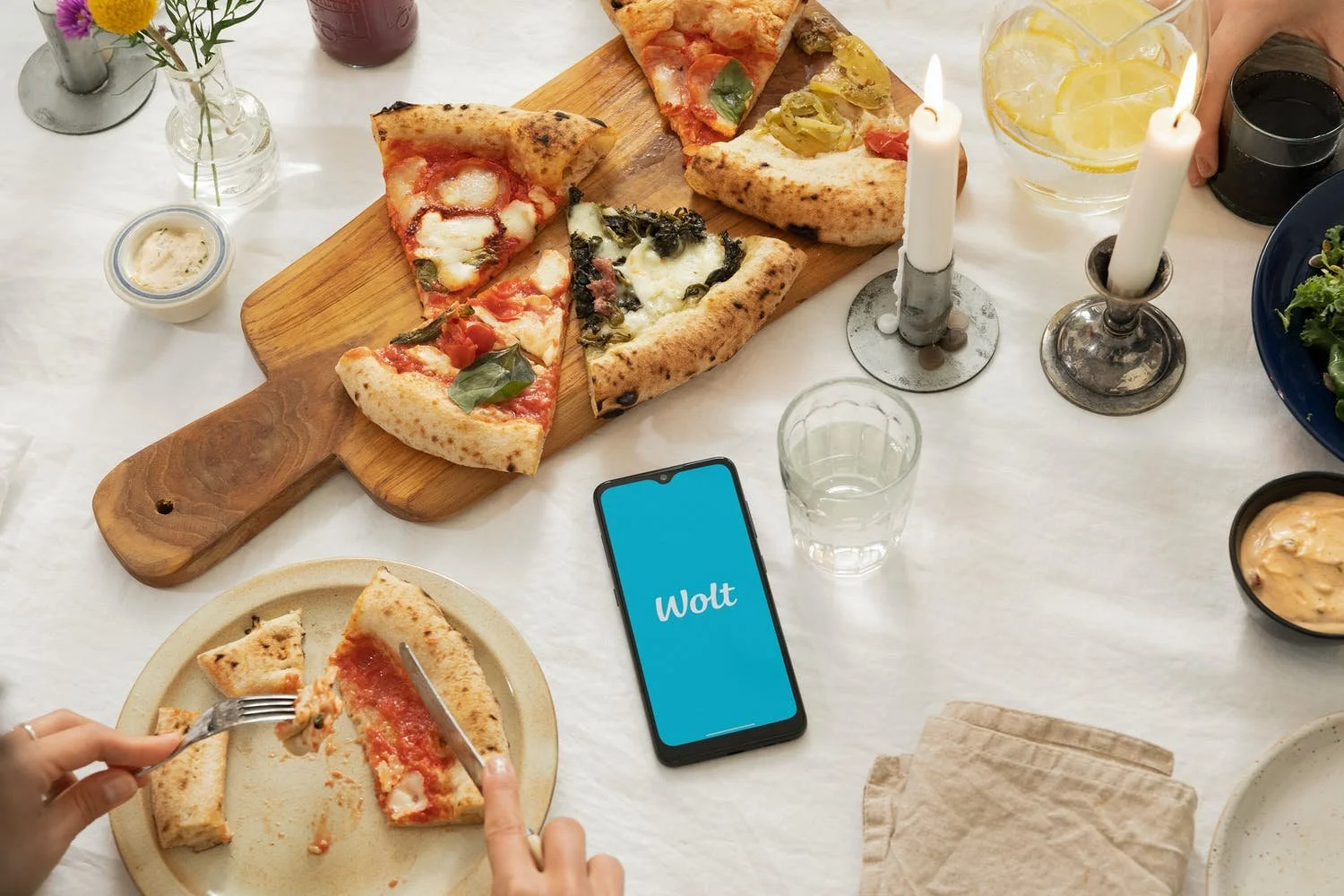 Pöydällä on pizza, juomaa ja puhelin, jossa lukee Wolt turkoosilla taustalla.