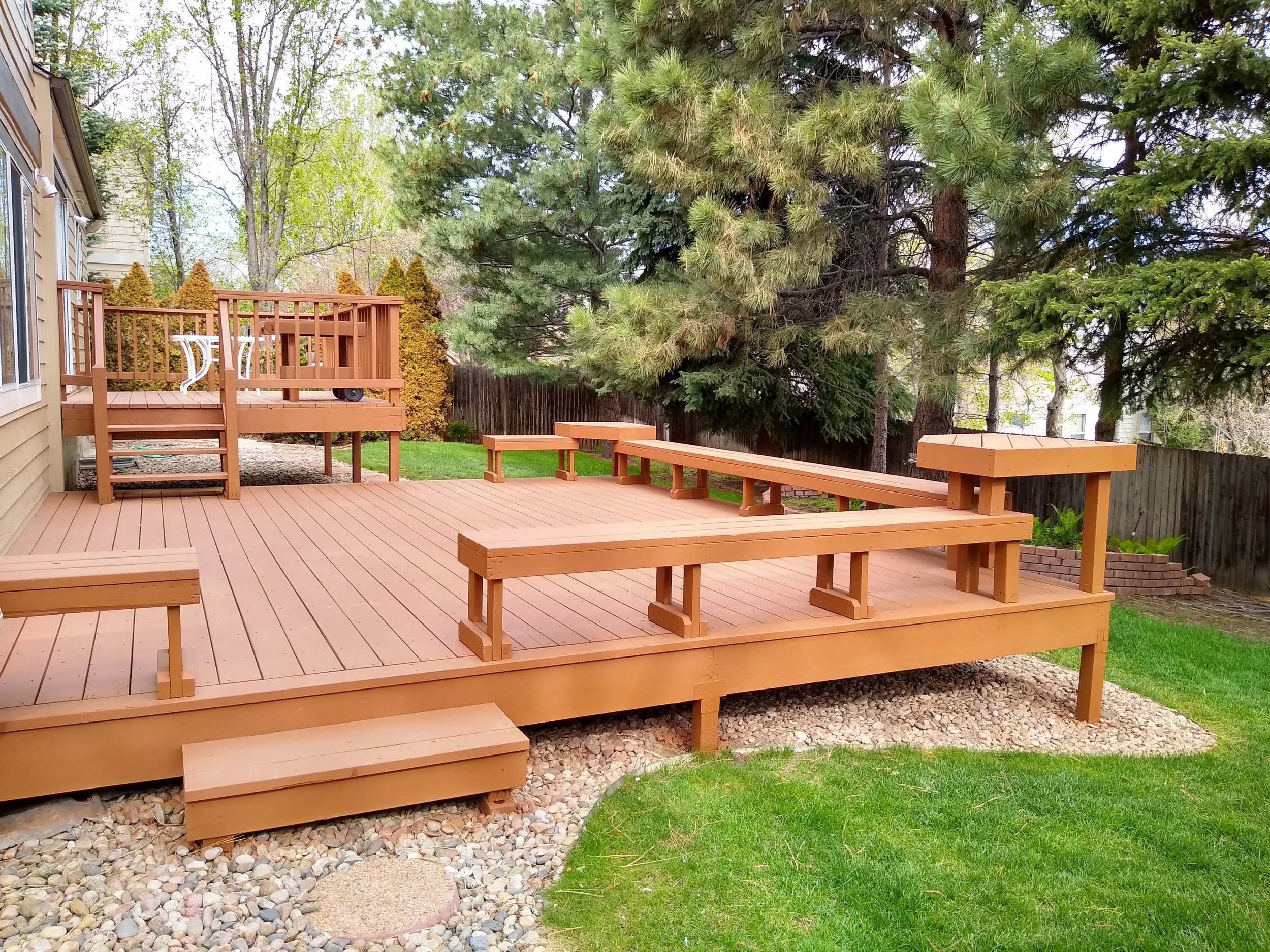 Wooden deck with built-in benches and stairs, surrounded by grass, rocks, and large trees in the backyard.