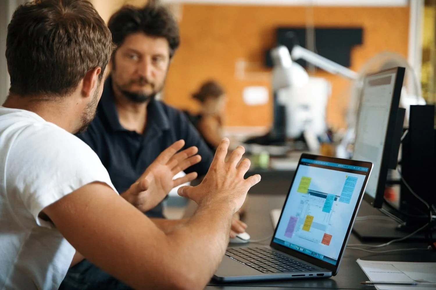 Due uomini siedono a un tavolo di lavoro, uno di fronte all'altro, mentre discutono. Sul tavolo c'è un laptop con un diagramma o un piano visivo visualizzato sullo schermo.