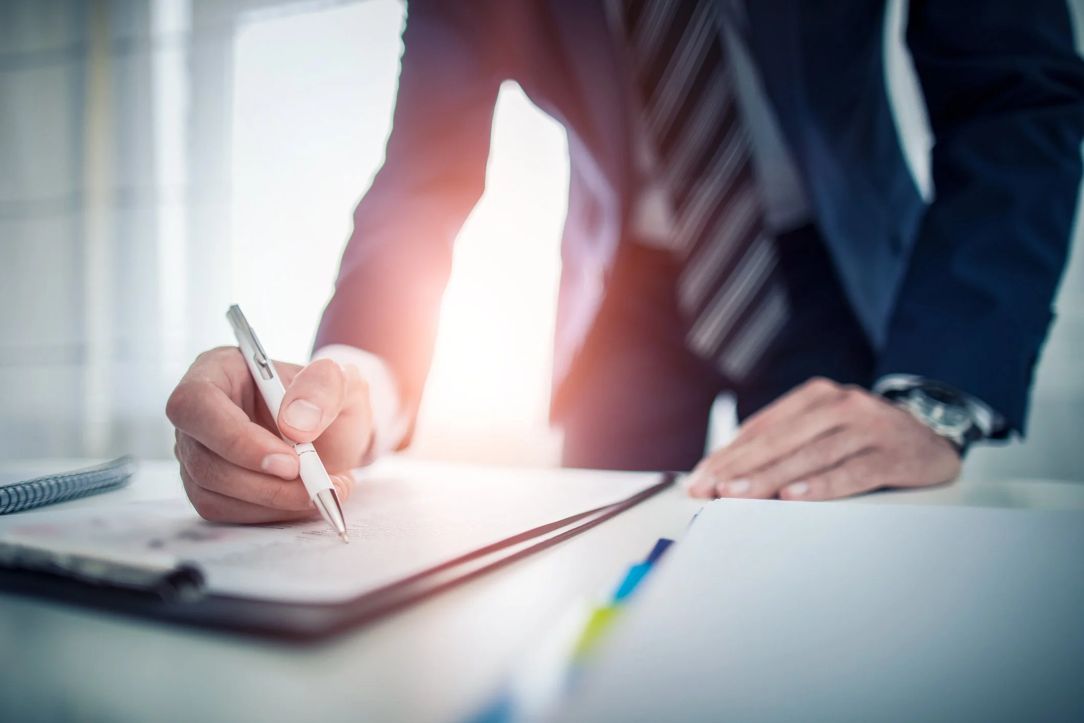 A person in a business suit signing a document on a white table, with sunlight coming through a window in the background.
