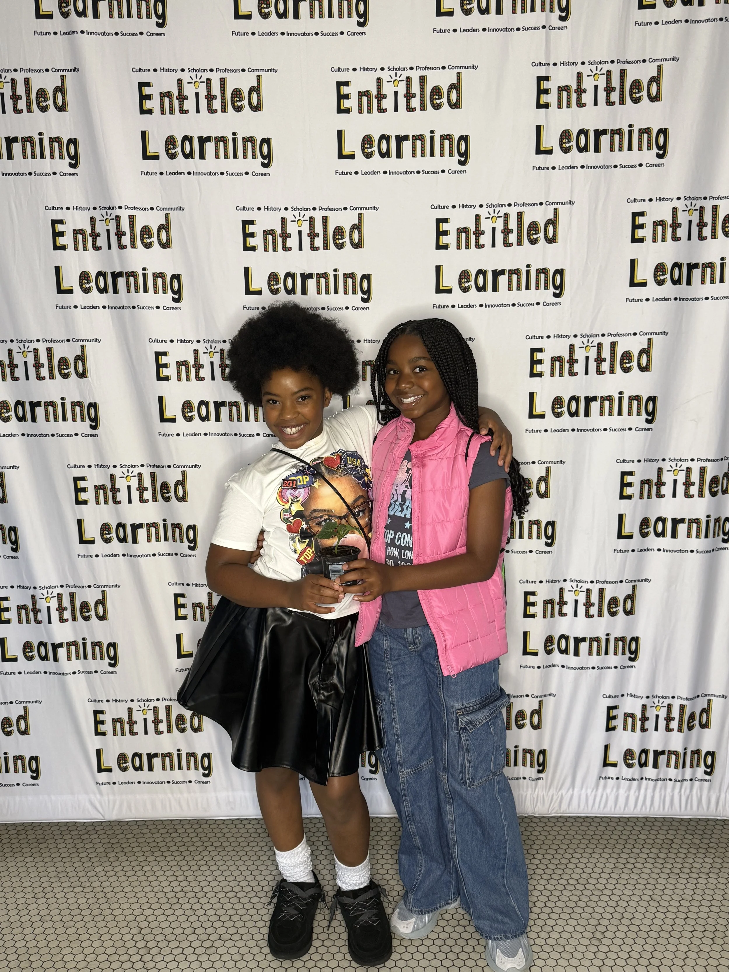 Two young girls smiling and posing together in front of a backdrop that reads "Entitled Learning." One girl, with curly afro hair, wearing a white T-shirt, black skirt, white socks, and black shoes, is holding a small potted plant. The other girl, with braided hair, wearing a pink vest over a gray T-shirt, denim cargo pants, and sneakers, has her arm around the girl with the plant.