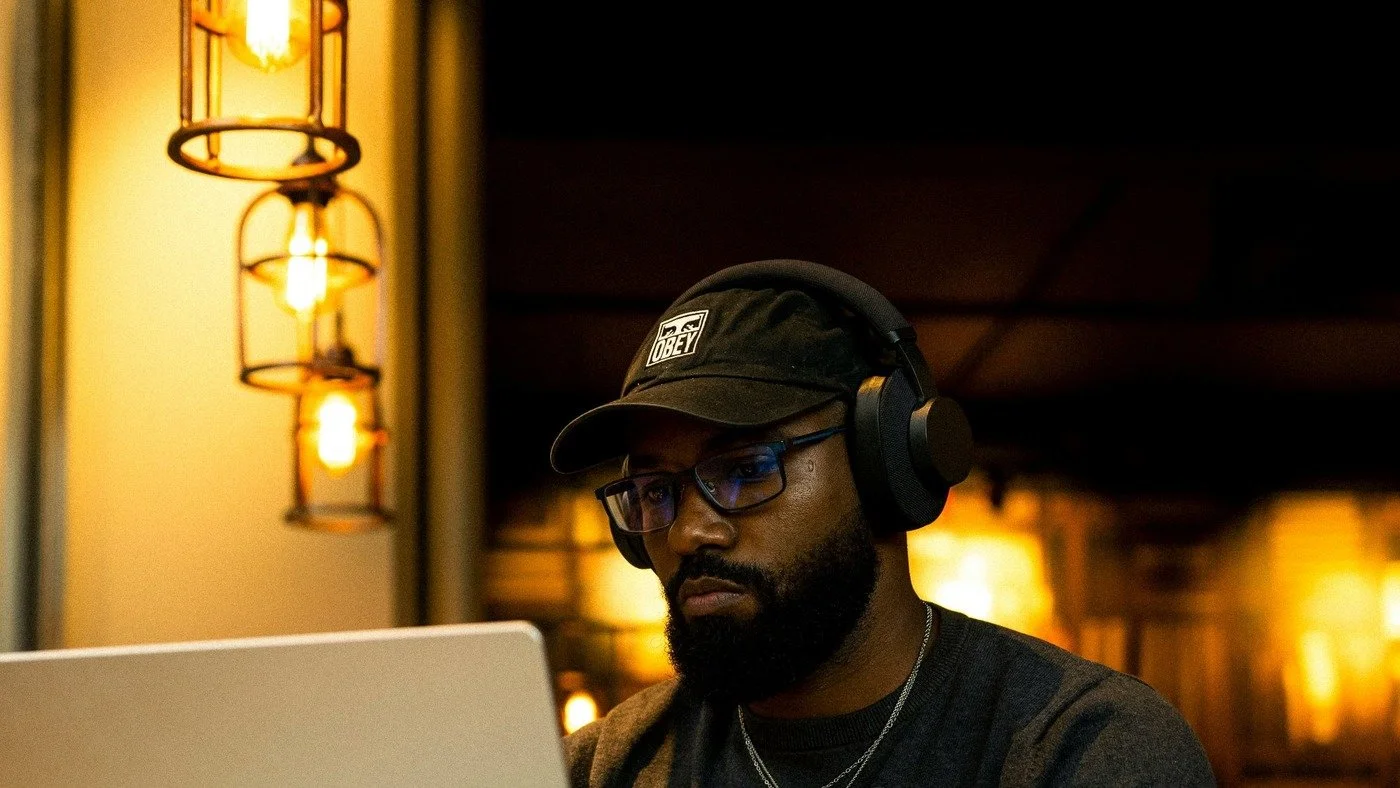 A young Black man in his mid-to-late twenties sits at a table working intently on a laptop. He wears over-ear headphones, glasses, and a dark cap. Warm pendant lights glow in the background.