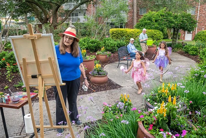 Two children dressed as fairies skipping along a sidewalk while their grandma paints a picture in the foreground.