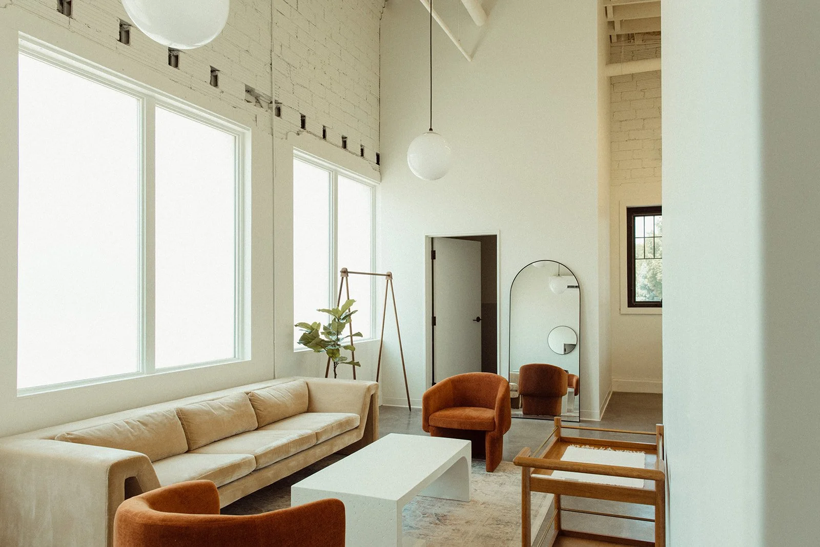 Modern living room with white walls, large windows, beige sofa, two orange armchairs, a white coffee table, a standing mirror, a small wooden bench, and a plant.