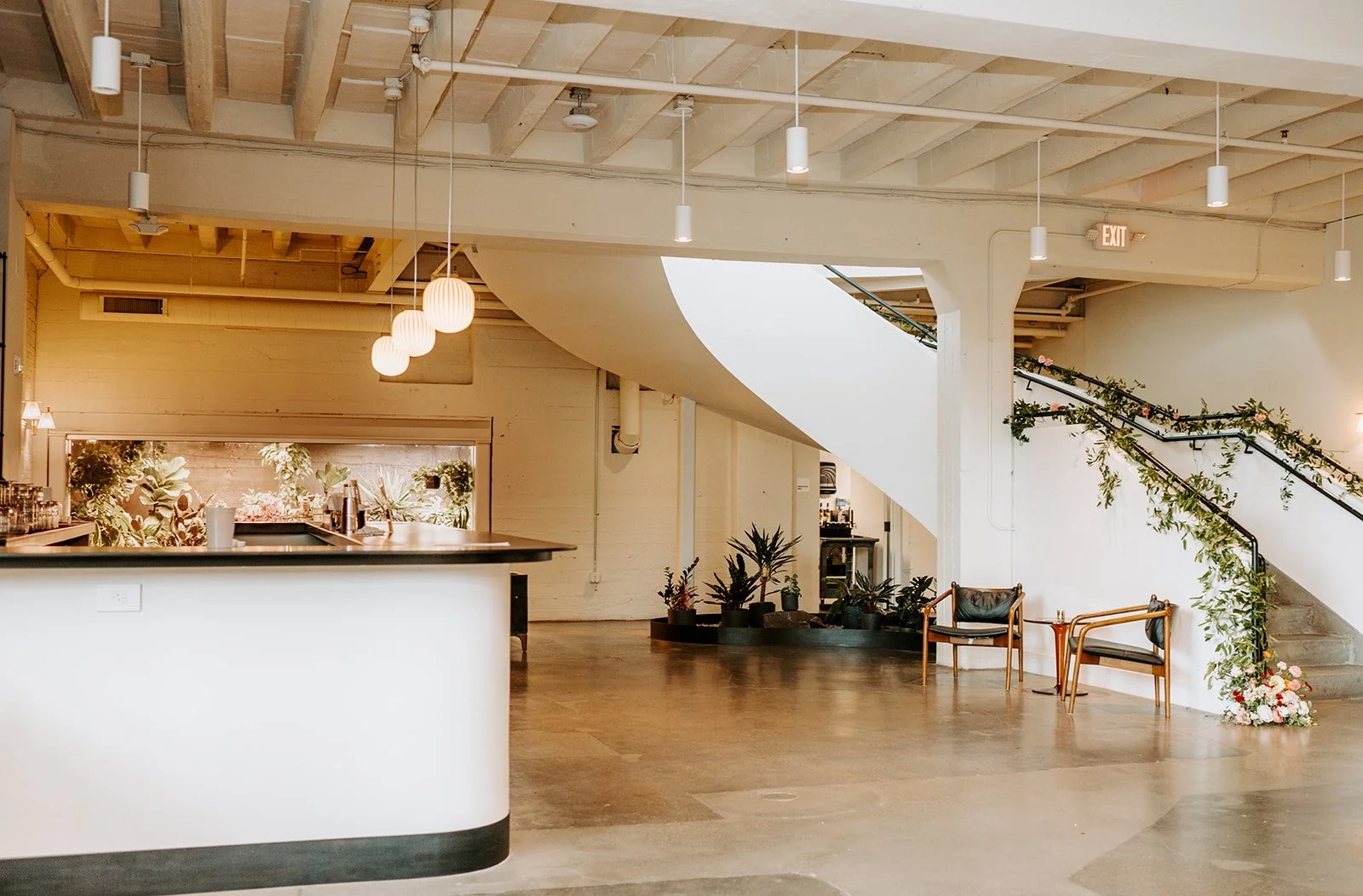 Empty interior space with a curved staircase decorated with green foliage and pink flowers, a small seating area with two wooden chairs and a table, a reception counter, and hanging pendant lights.