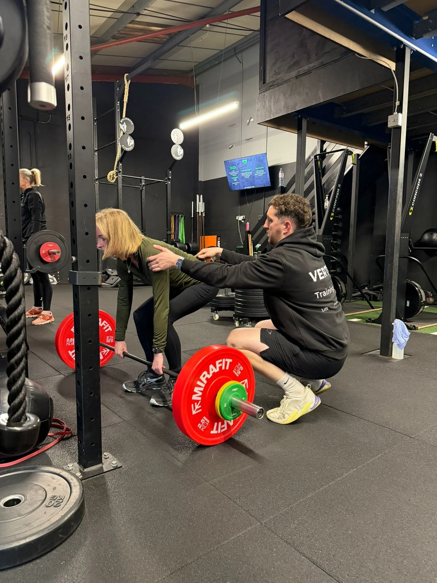 Two people in a gym, one woman is doing a deadlift with a barbell loaded with red weights, while a man appears to be coaching or assisting her.