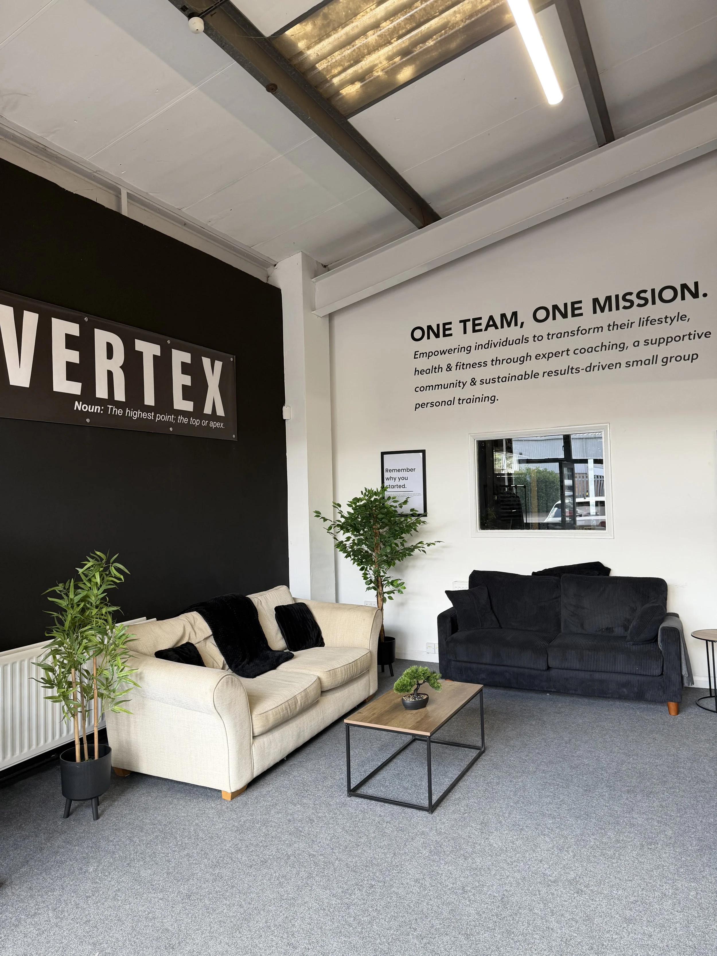 Modern office lounge with beige and black sofas, a small wooden coffee table, potted plants, and motivational wall posters. The room features gray carpet flooring, a window, and a white wall with a large black and white wall art.