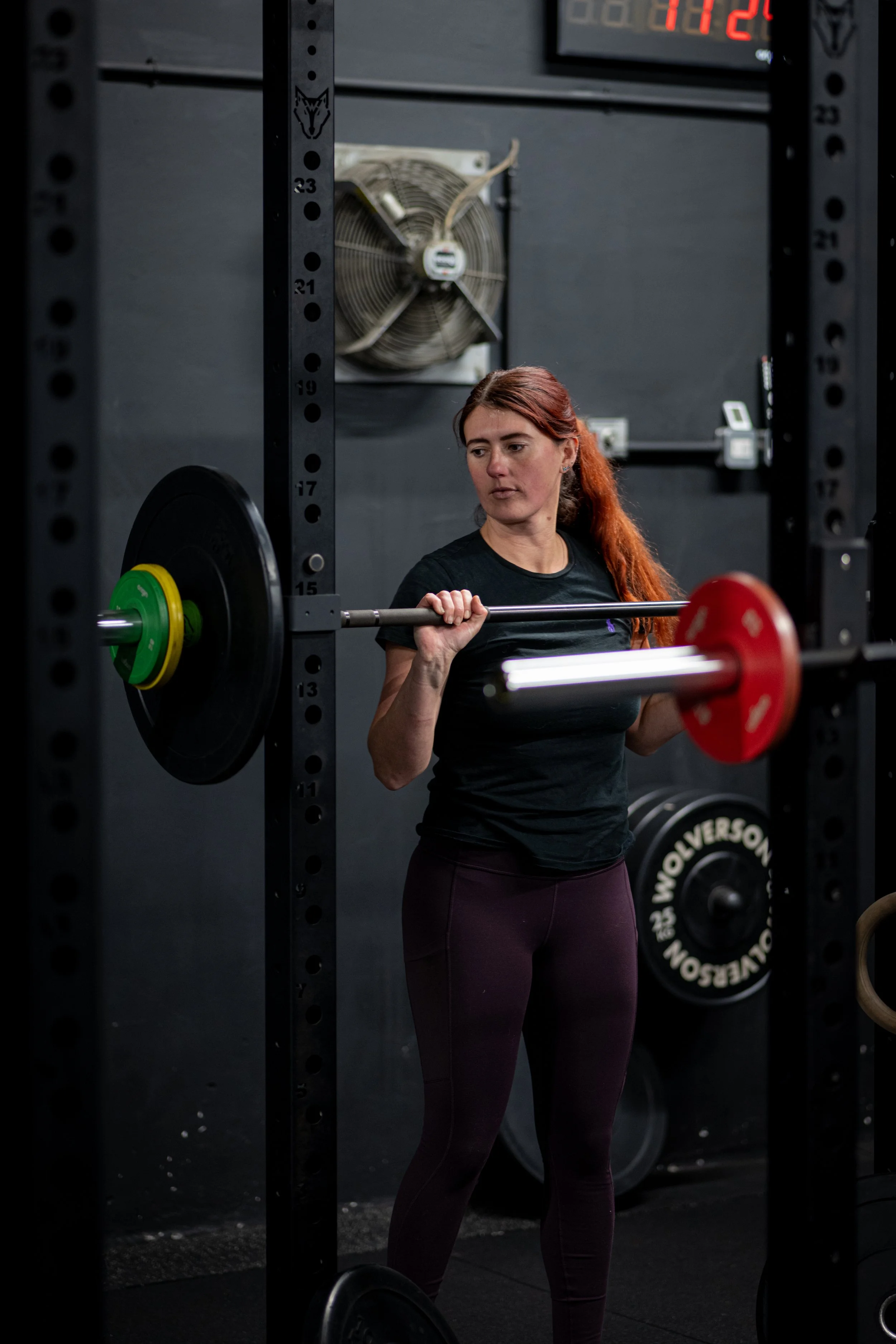 A woman with long red hair, wearing a black athletic t-shirt and dark purple leggings, is lifting a barbell with weights at a gym. She is standing inside a squat rack with a fan on the wall behind her and a digital clock above.