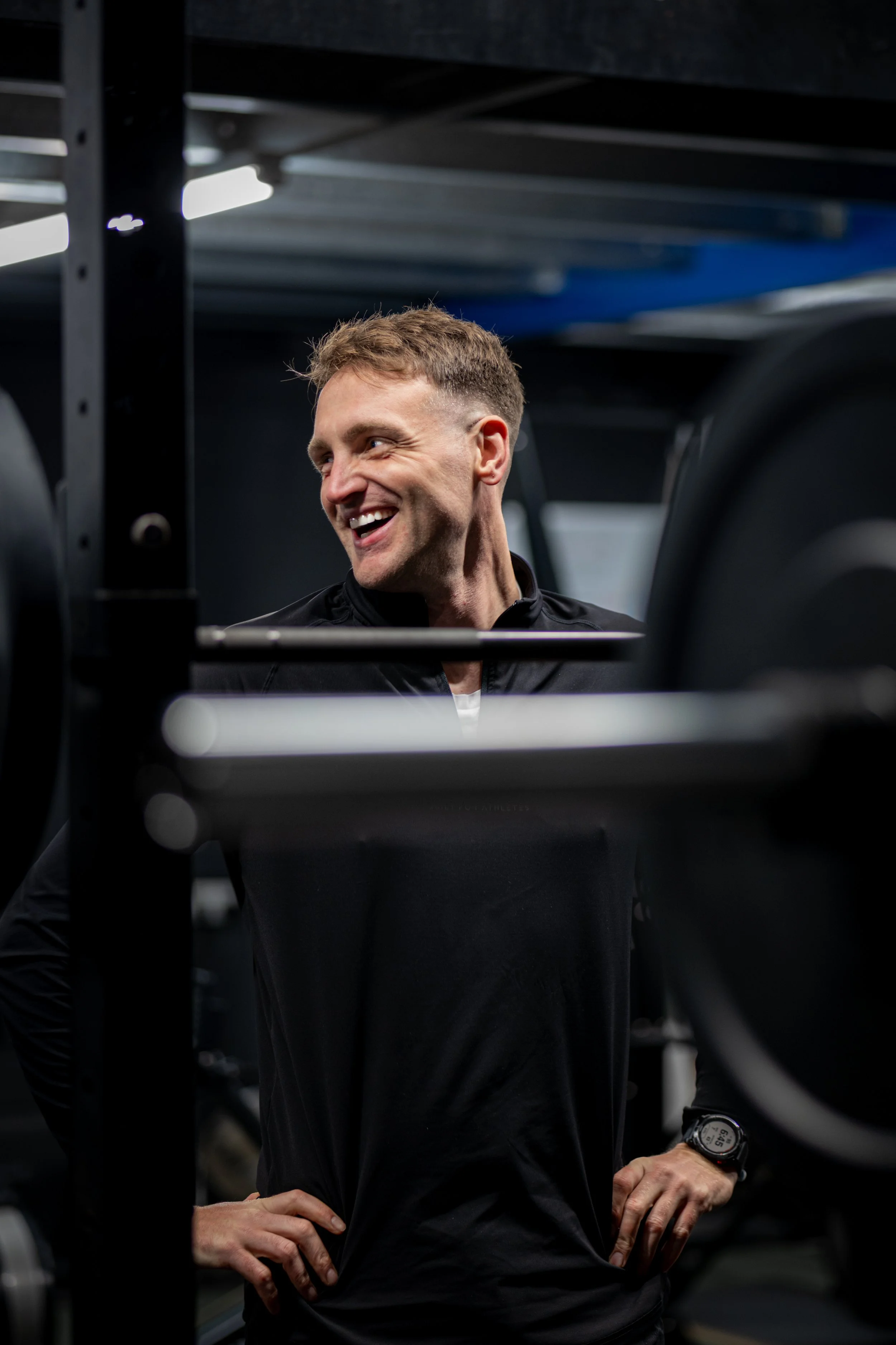 A man smiling in a gym, wearing a black workout jacket and a watch, with gym equipment in the background.
