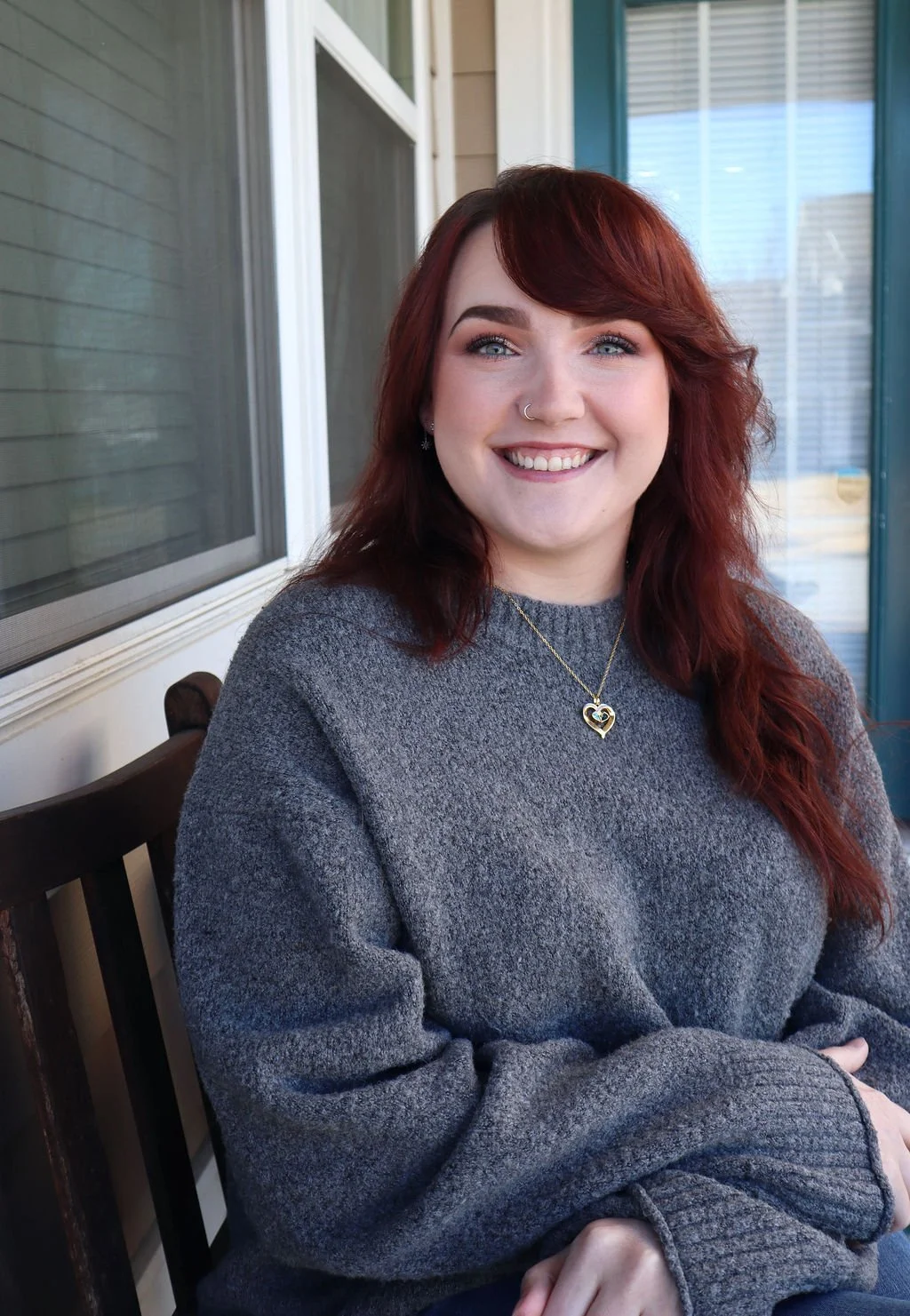 An image of Holly who is a white person with long red hair, wearing a gray sweater and gold necklace. They sit outside.