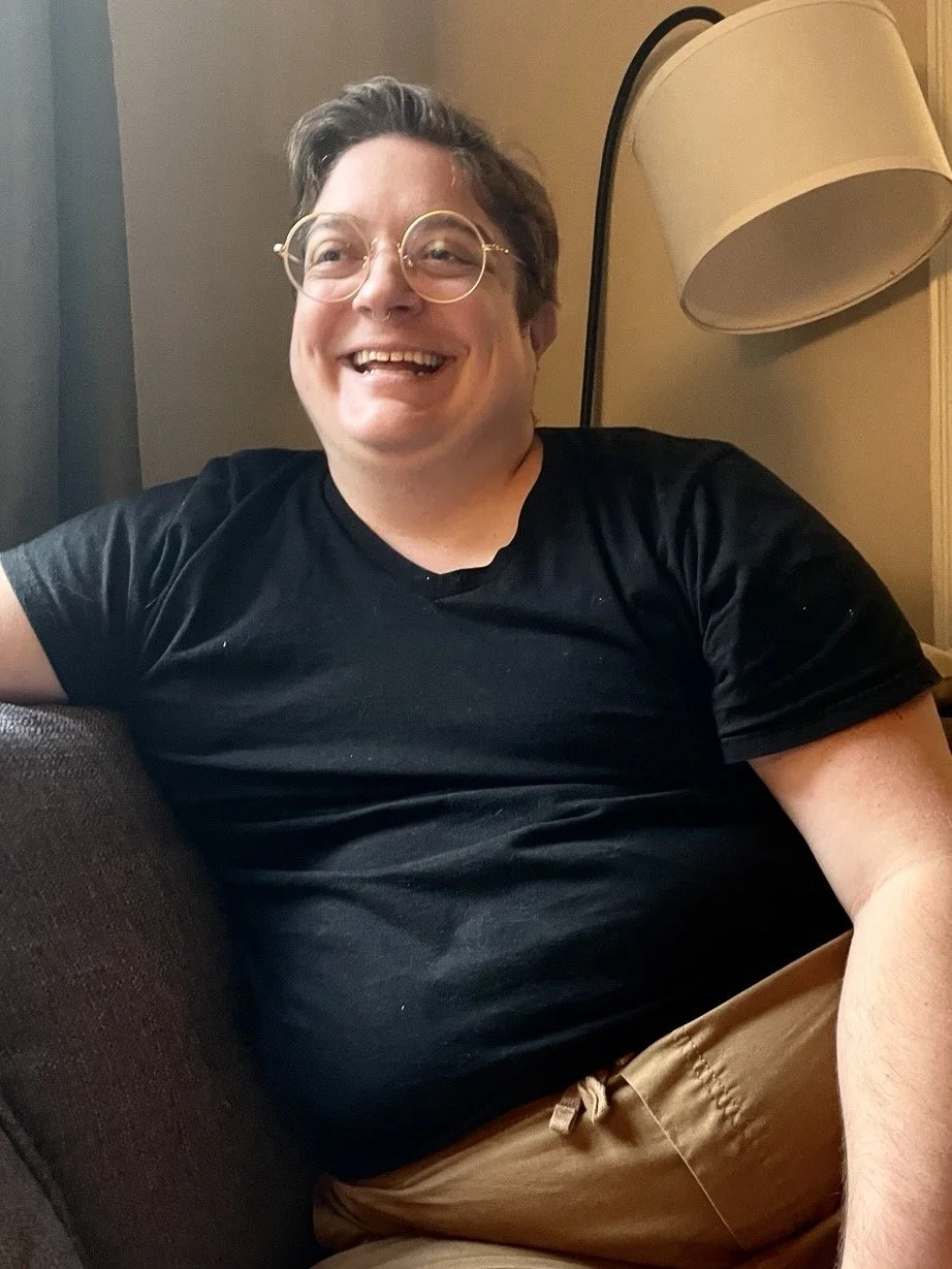 A picture of AJ who is a white male wearing glasses, a black t-shirt and sitting on a couch. Short hair and he smiles