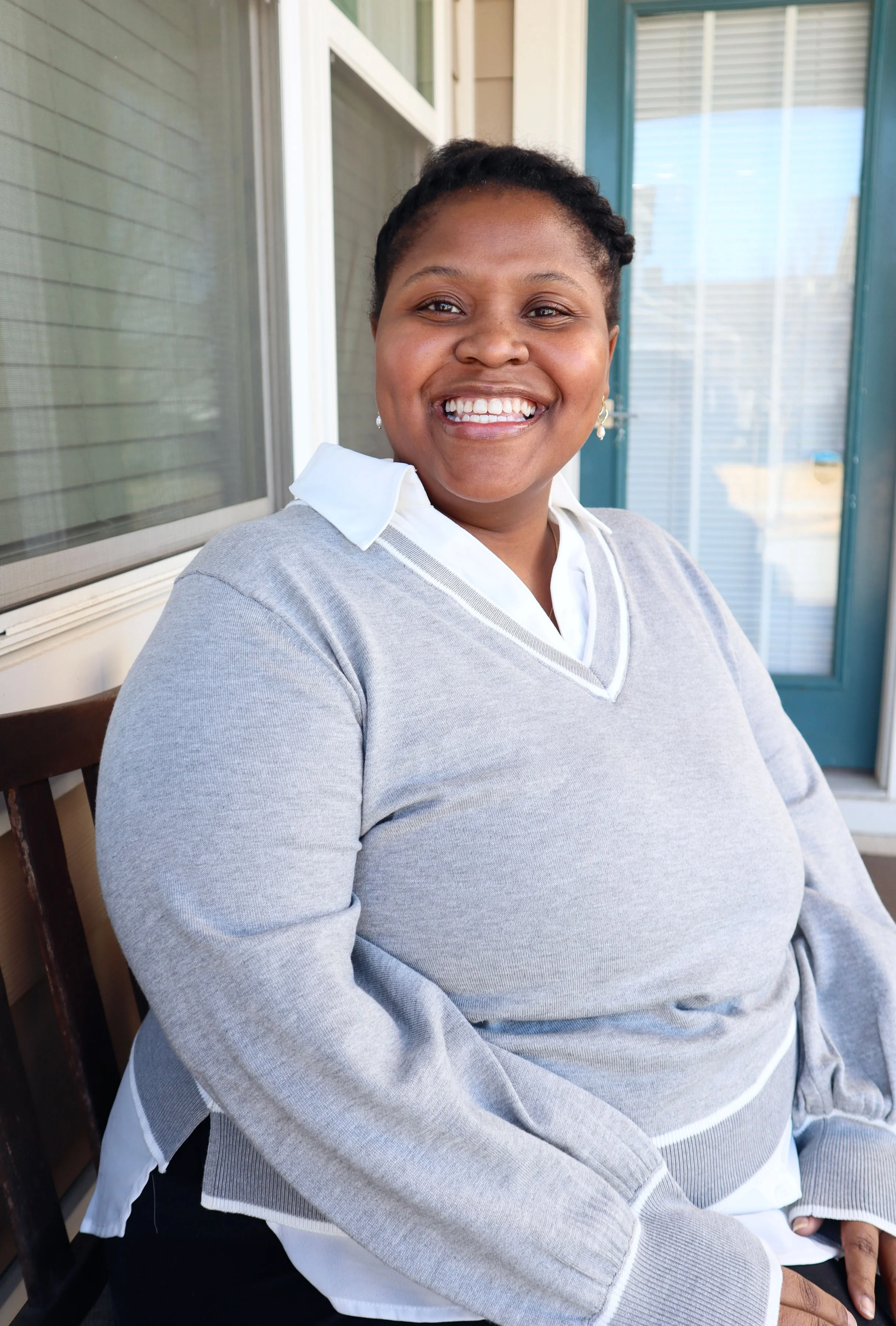 Najjiya is a black woman wearing a gray sweater with a white collared shirt underneath, smiling at the camera