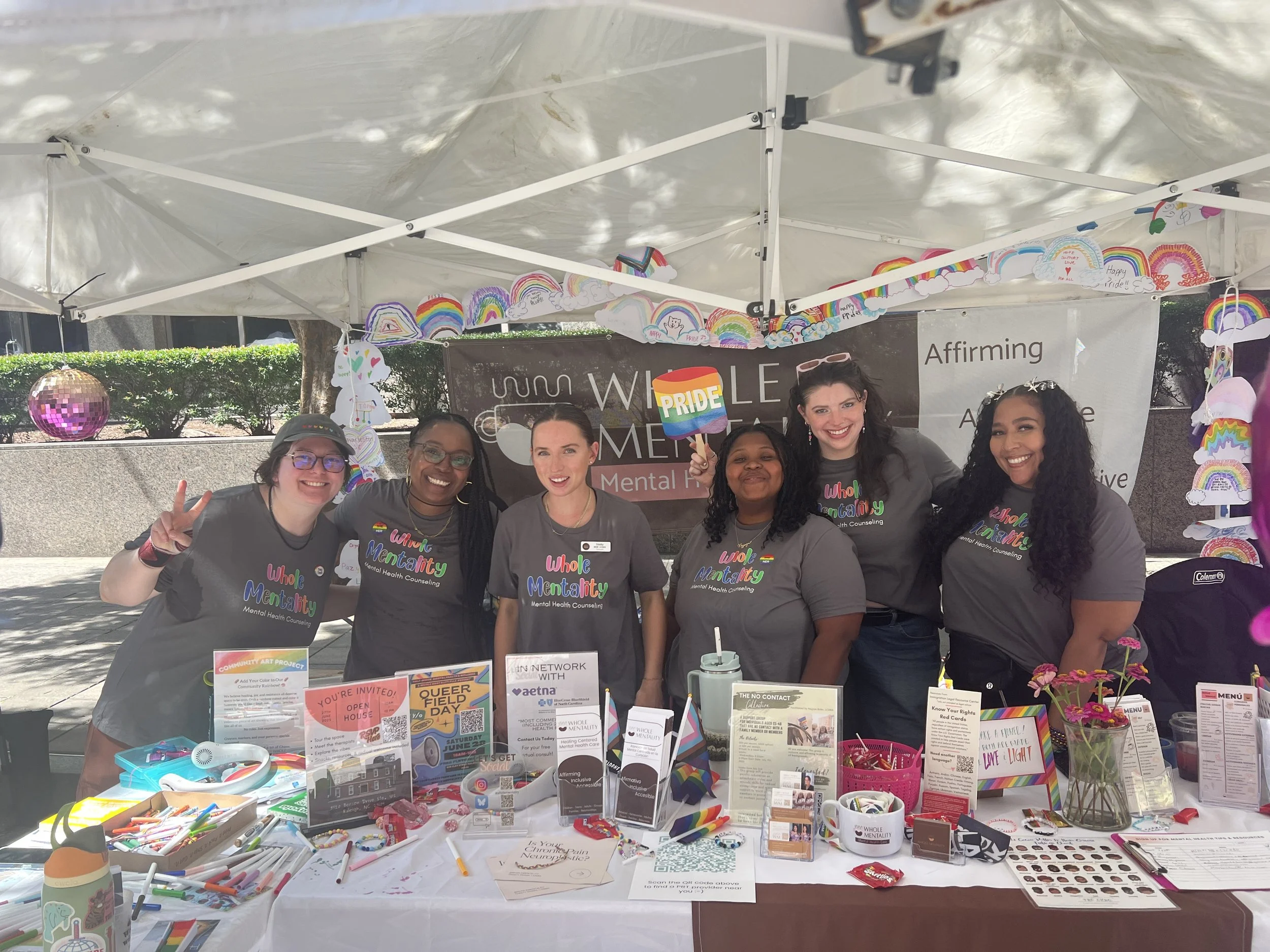 An image of the whole Mentality team at their PRIDE table filled with information, brochures, candy, and goodies. The entire team wears the same Whole Mentality shirt in front of a Whole entality sign. 