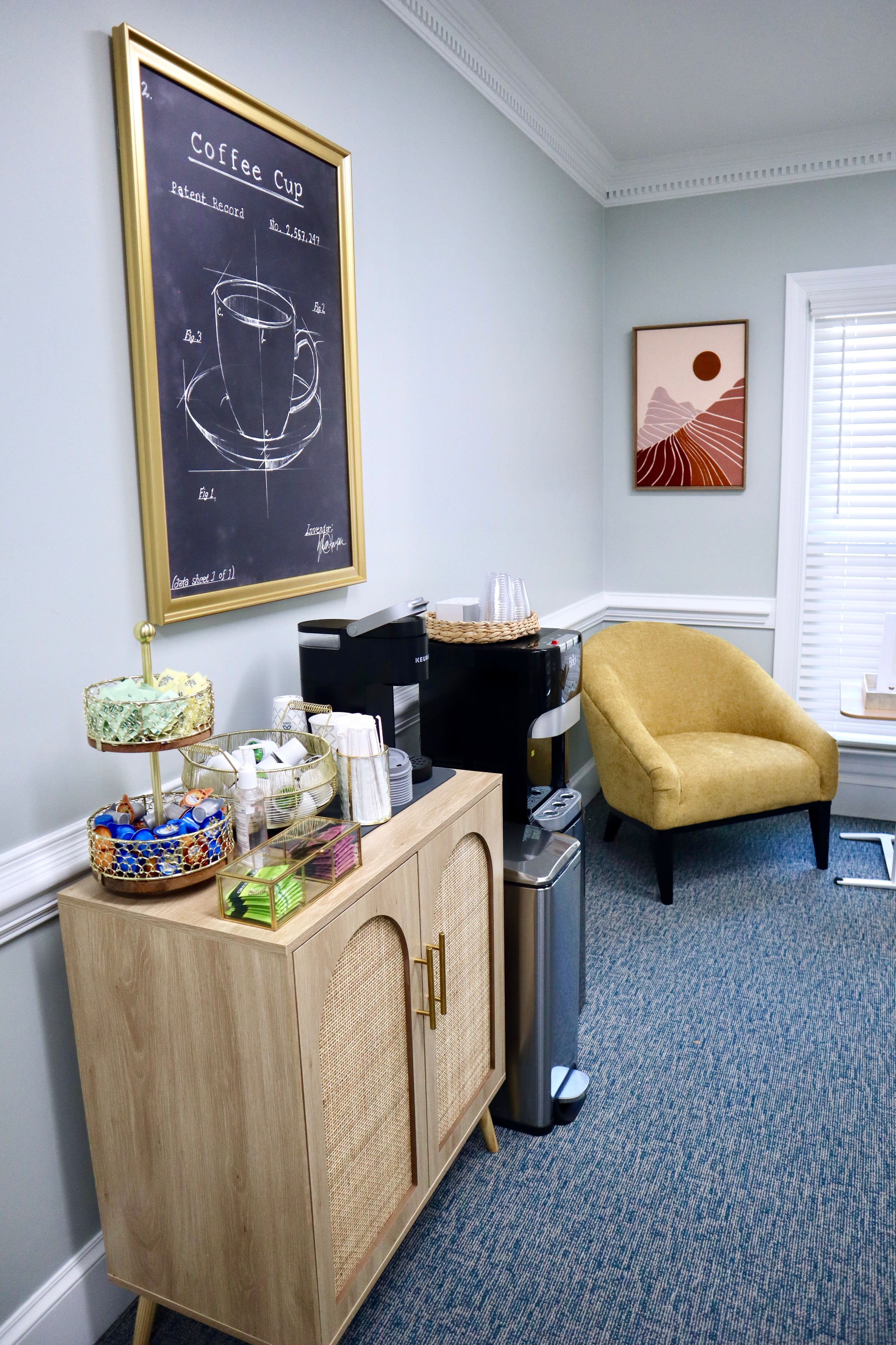 A picture of the coffee station from a different angle. Next to the coffee station is a water filter station. The yellow chair is in the corner under colorful artowrk .