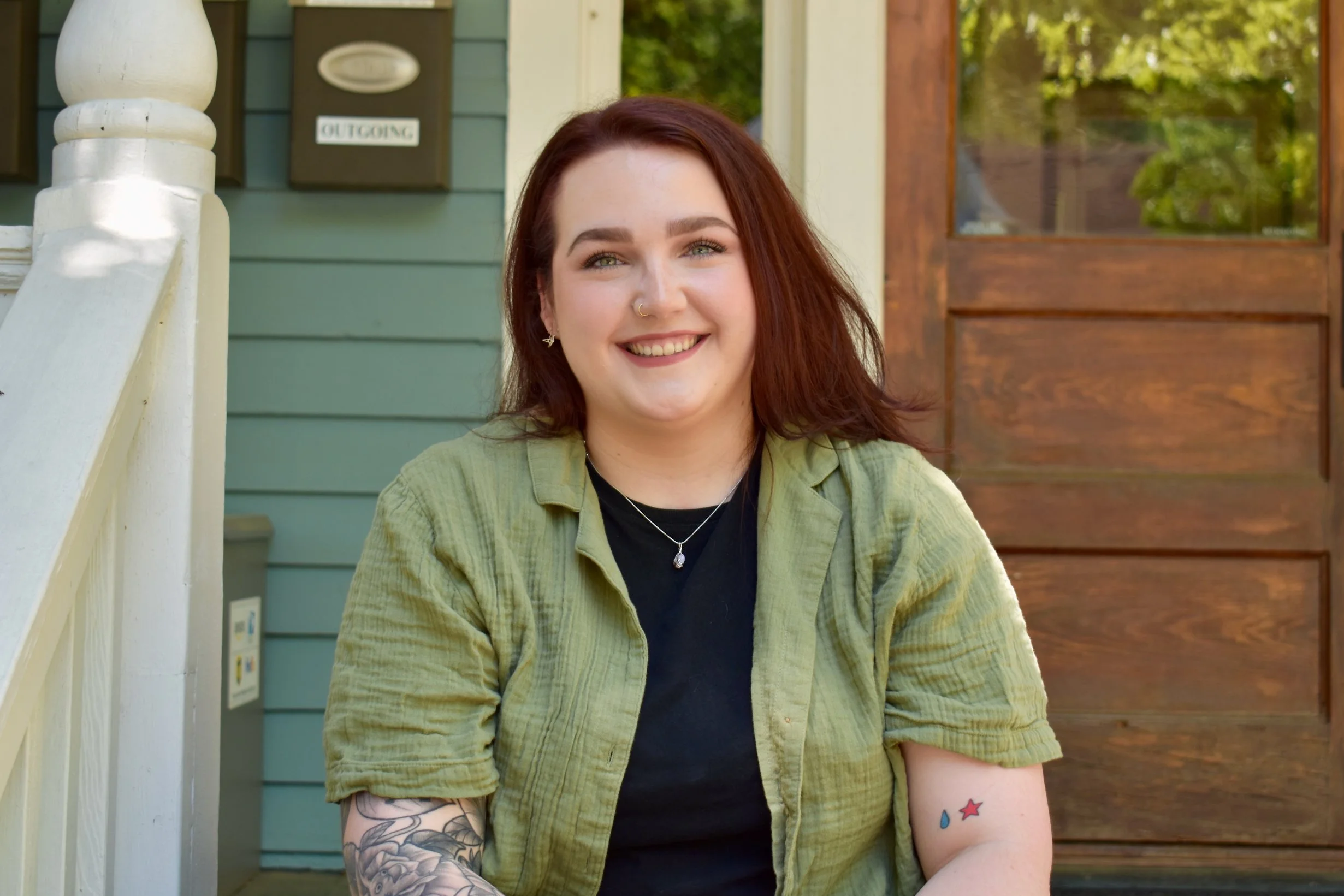 An image of Haley who is a white person with long red heair, wearing a green blouse with a black shirt underneath. They sit outside and you can see parts of their tattooed arms