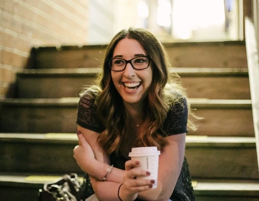 An image of Eden, a white female, sitting on a staircase, smiling and holding a cup of coffee. Eden wears glasses and a black top.